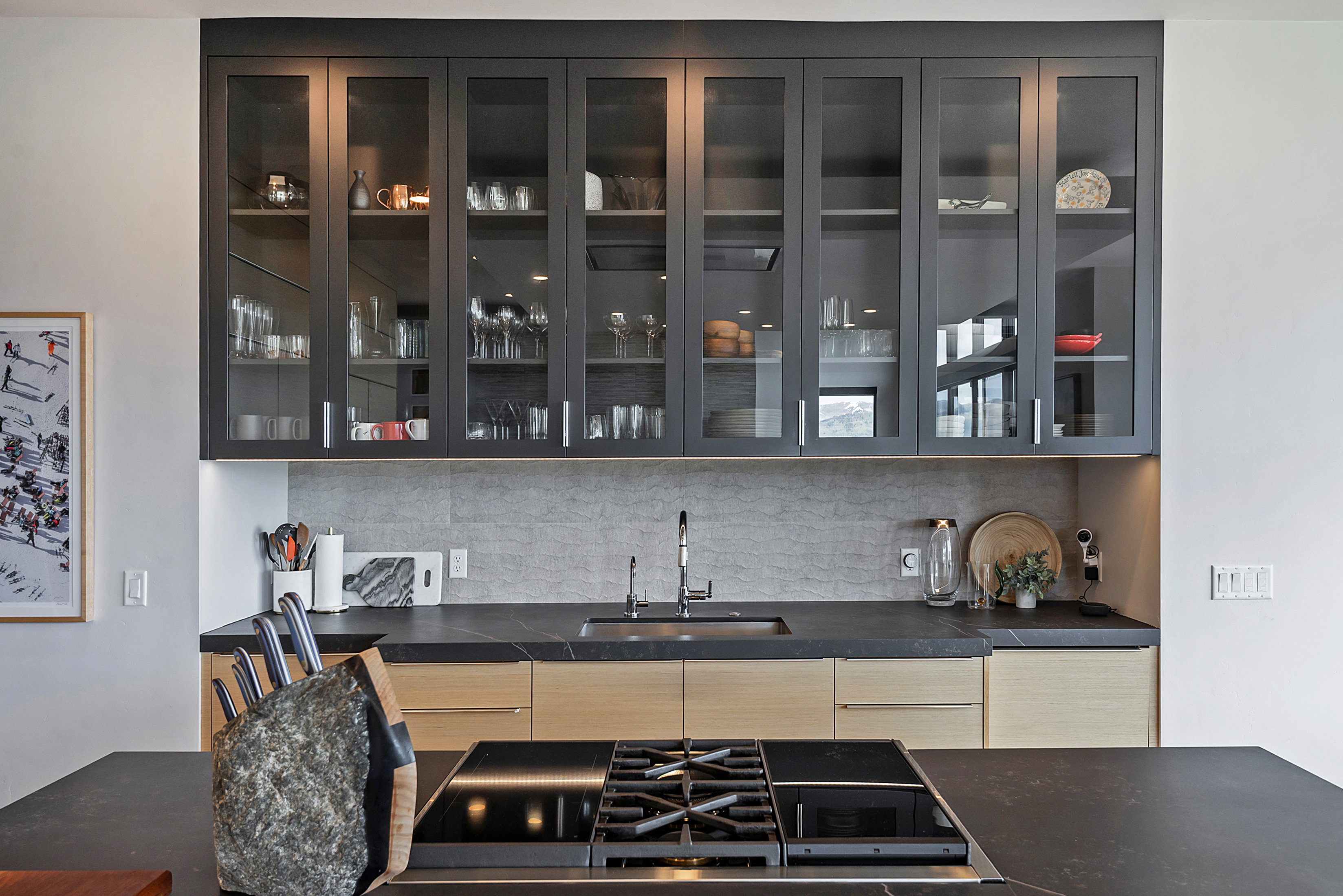 Sleek kitchen featuring black frame glass-front upper cabinetry with integrated lighting, wood-finish lower cabinets, a black countertop with an integrated sink, a gray textured backsplash, and a black cooktop