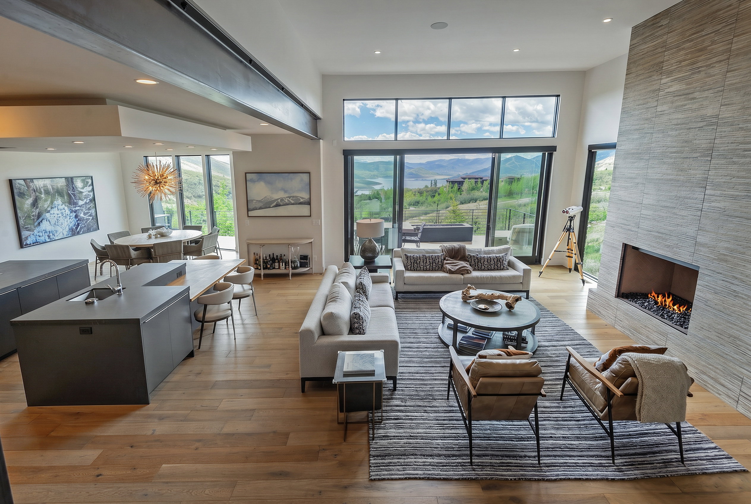Spacious living area featuring expansive floor-to-ceiling windows, wood-finish flooring, a contemporary fireplace with a stone surround, and an open-concept kitchen island with seating