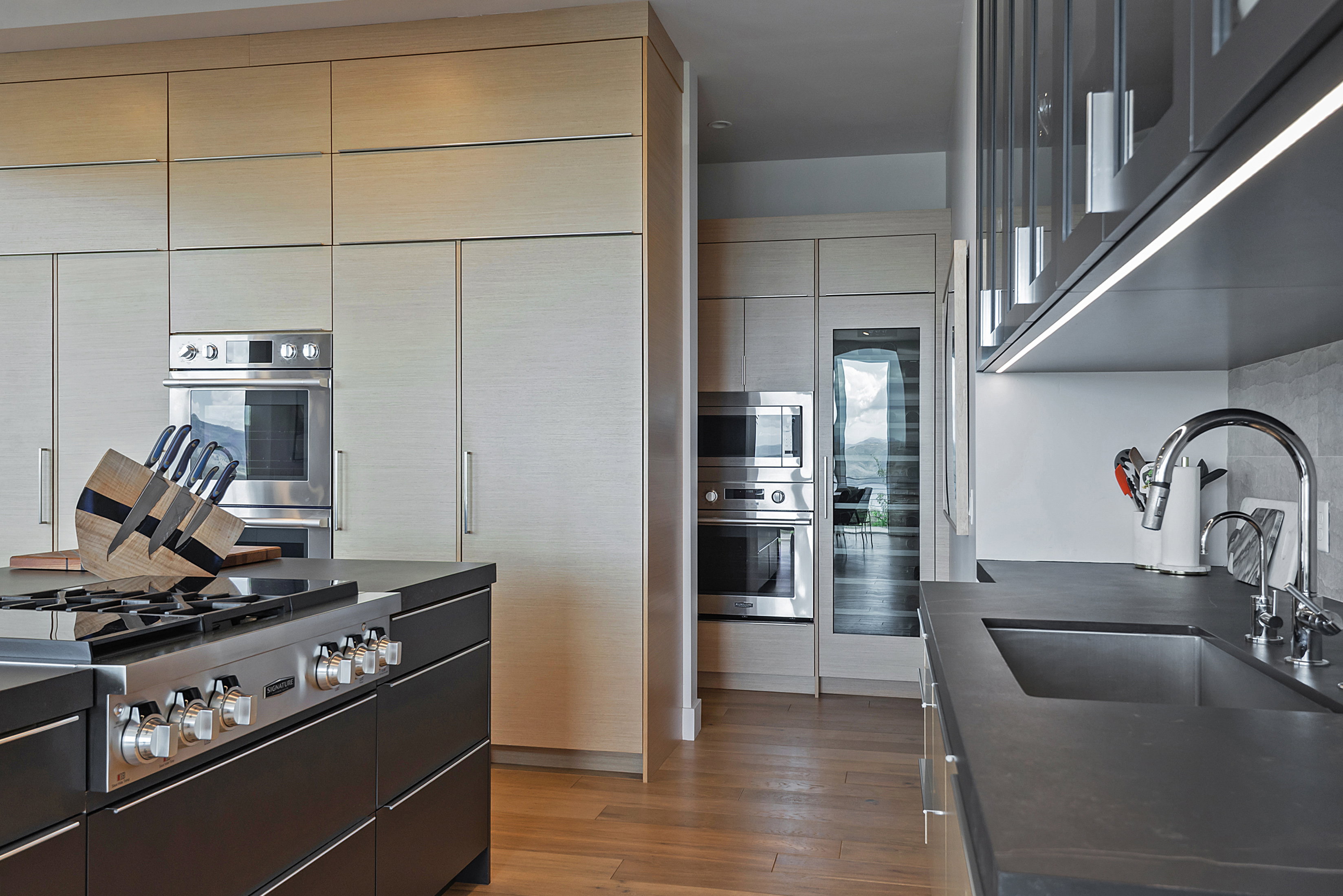 Modern kitchen featuring a large island with a gas range, integrated stainless steel appliances, light wood-finish cabinetry, dark countertops, and wood-finish flooring
