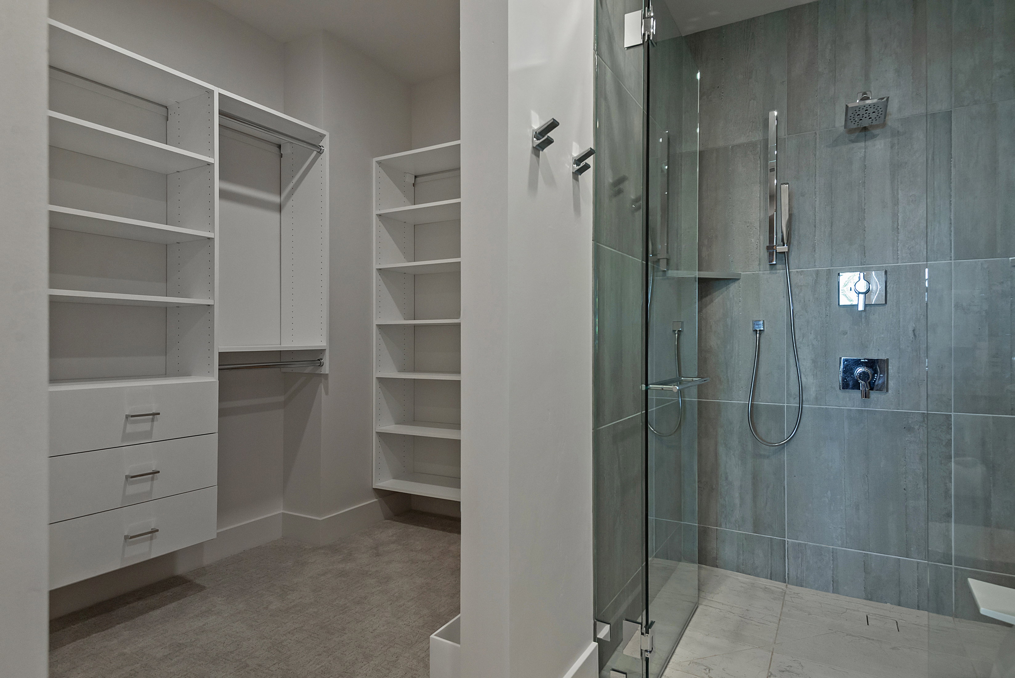 Walk-in closet with custom white built-in shelving and drawers, alongside a glass-enclosed shower featuring dark grey tile walls, a rain showerhead, and a handheld shower attachment