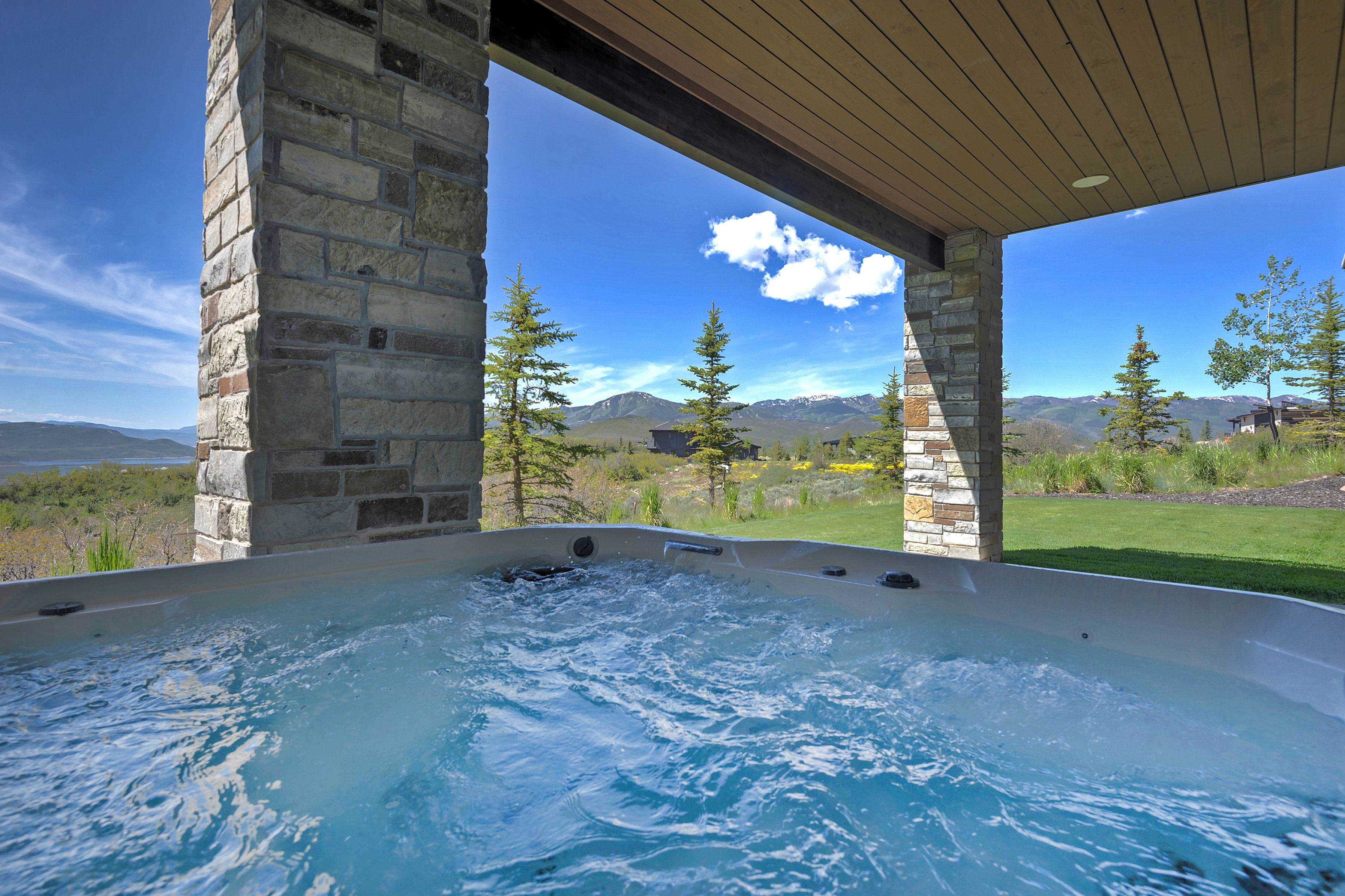 Outdoor spa situated under a covered patio with stone columns, featuring a clear sky, distant mountains, and a grassy area
