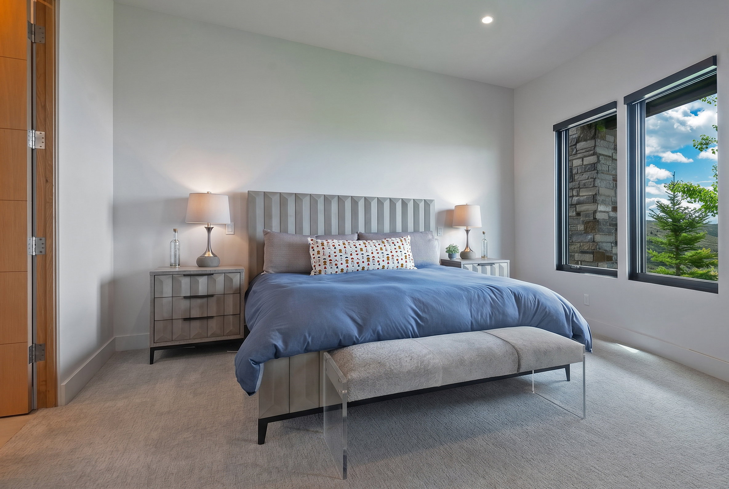 Carpeted bedroom featuring two large windows with dark trim, offering natural light and glimpses of outdoor scenery