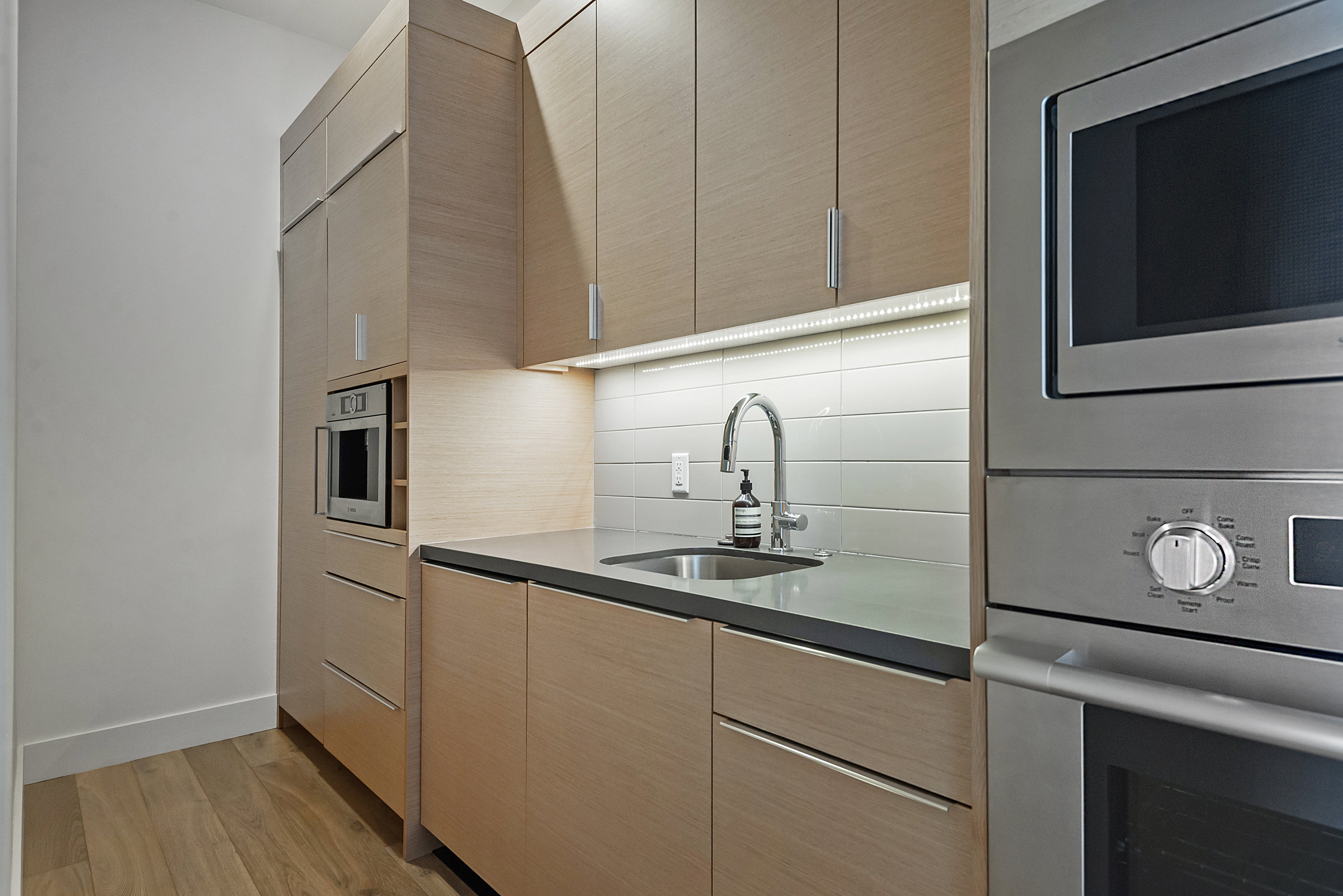 Streamlined kitchen area featuring wood-finish cabinetry with integrated appliances, stainless steel sink, and light-toned horizontal tile backsplash