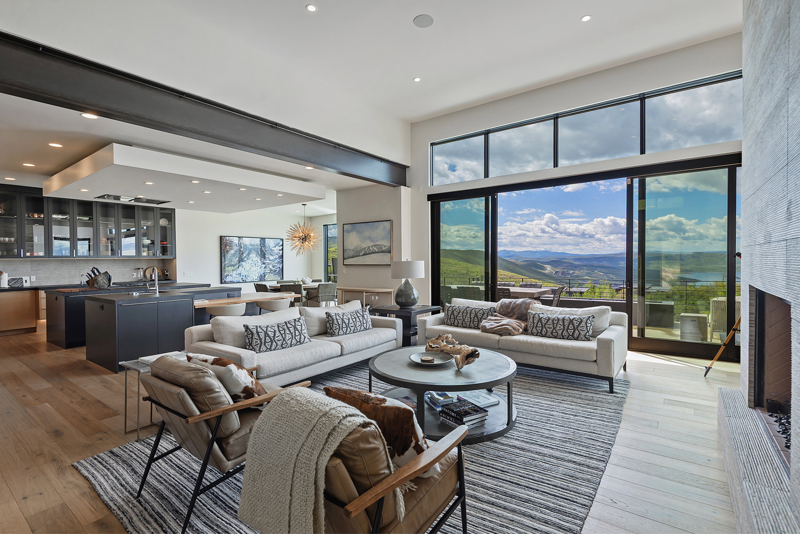 Expansive open-concept living area featuring a wood-finish flooring, a contemporary fireplace with a stone surround, and floor-to-ceiling sliding glass doors offering panoramic mountain and lake scenery