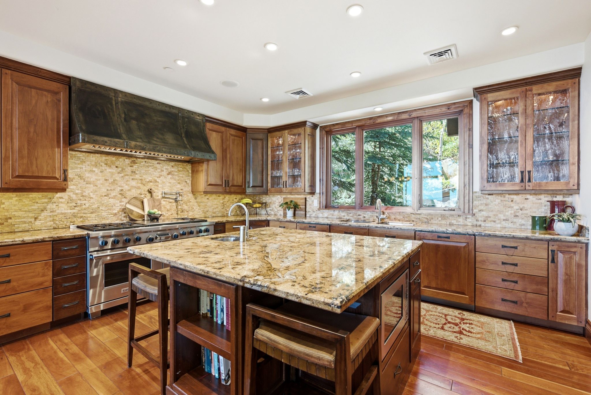 Gourmet kitchen featuring a large granite-topped island, a professional-grade range with an industrial-style hood, wood-finish cabinetry with some glass-front doors, a stone tile backsplash, and a window overlooking exterior foliage