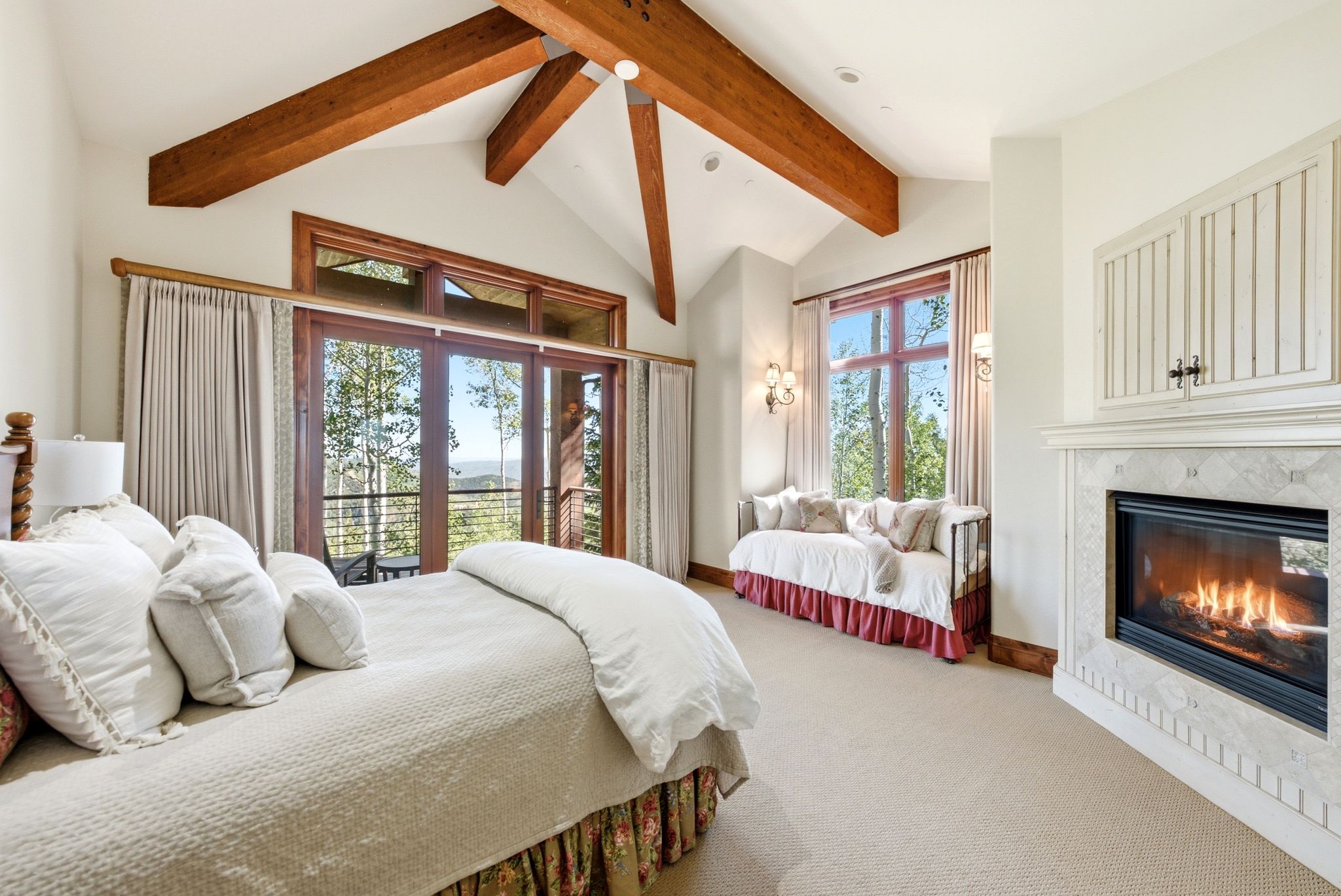 Vaulted ceiling with exposed wood beams, large glass doors opening to a balcony, built-in fireplace with a tile surround, built-in wall sconces, and a window seat