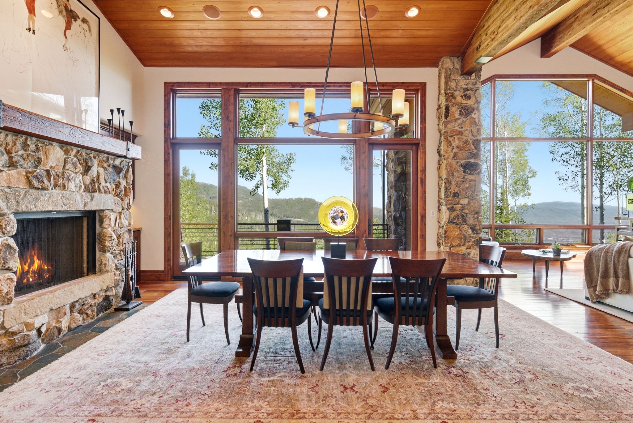 Dining area featuring extensive wood-frame windows, a stone fireplace, wood-finish flooring, a wood beam ceiling, and a circular chandelier
