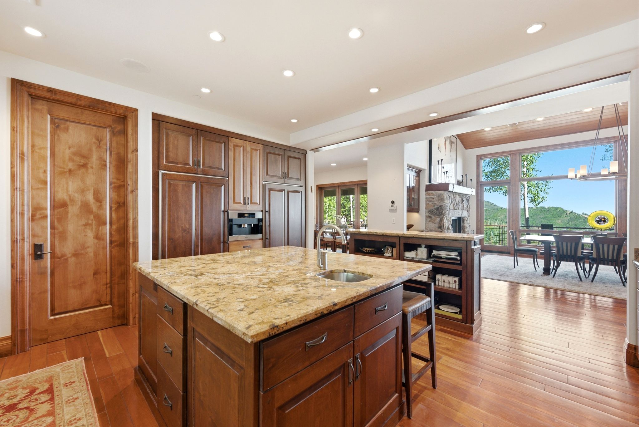 Spacious kitchen featuring a large central island with an undermount sink and granite countertop, rich wood-finish flooring, integrated appliances, and recessed lighting