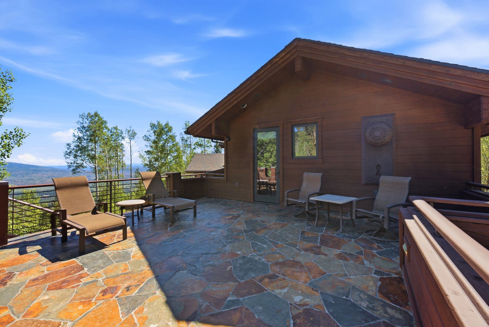 Expansive stone patio featuring a horizontal cable railing system, a wood-sided exterior, and mature trees in the background