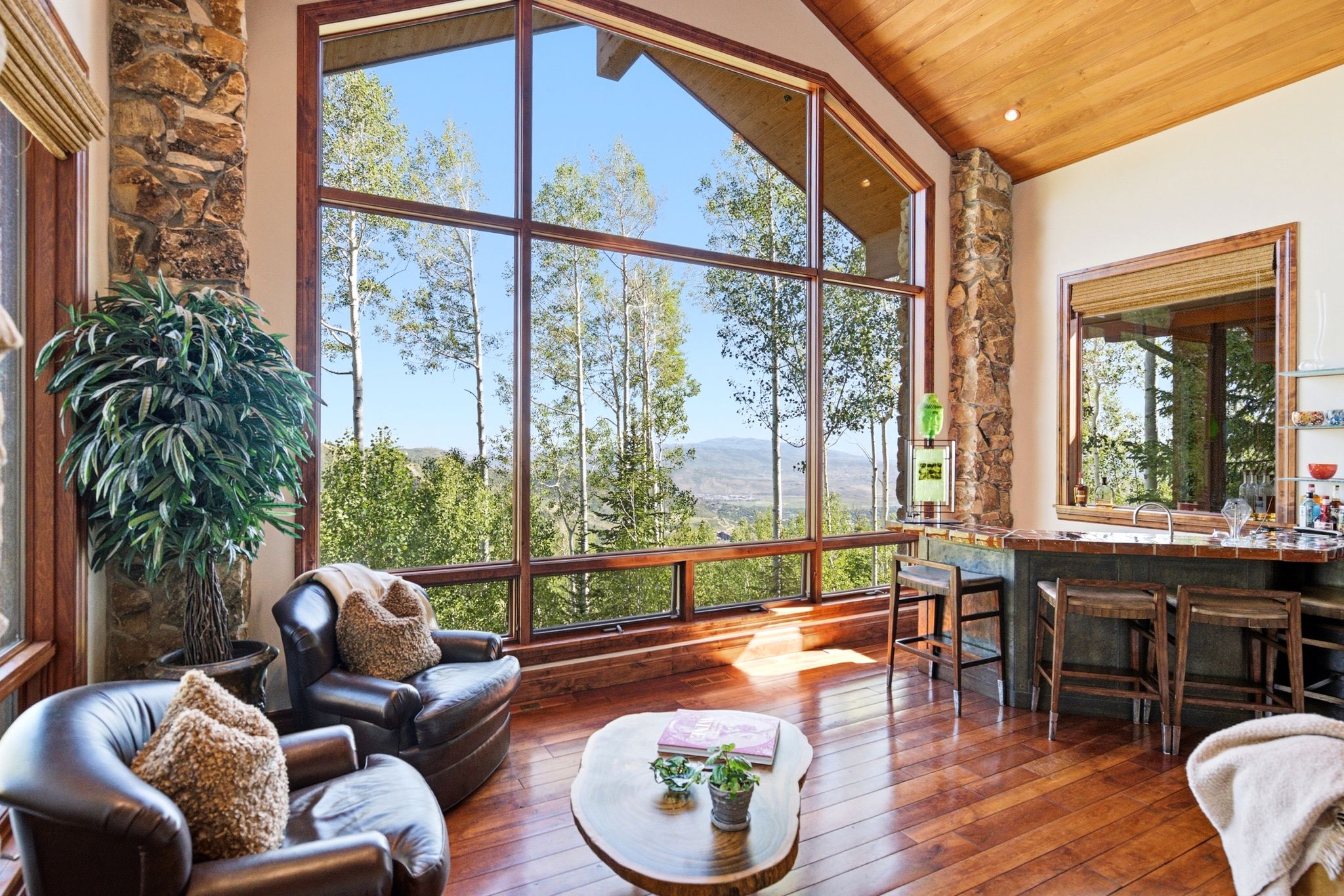 Great room featuring expansive windows with mountain and tree scenery, vaulted wood-paneled ceilings, rich wood-finish flooring, and a built-in wet bar with a tiled countertop