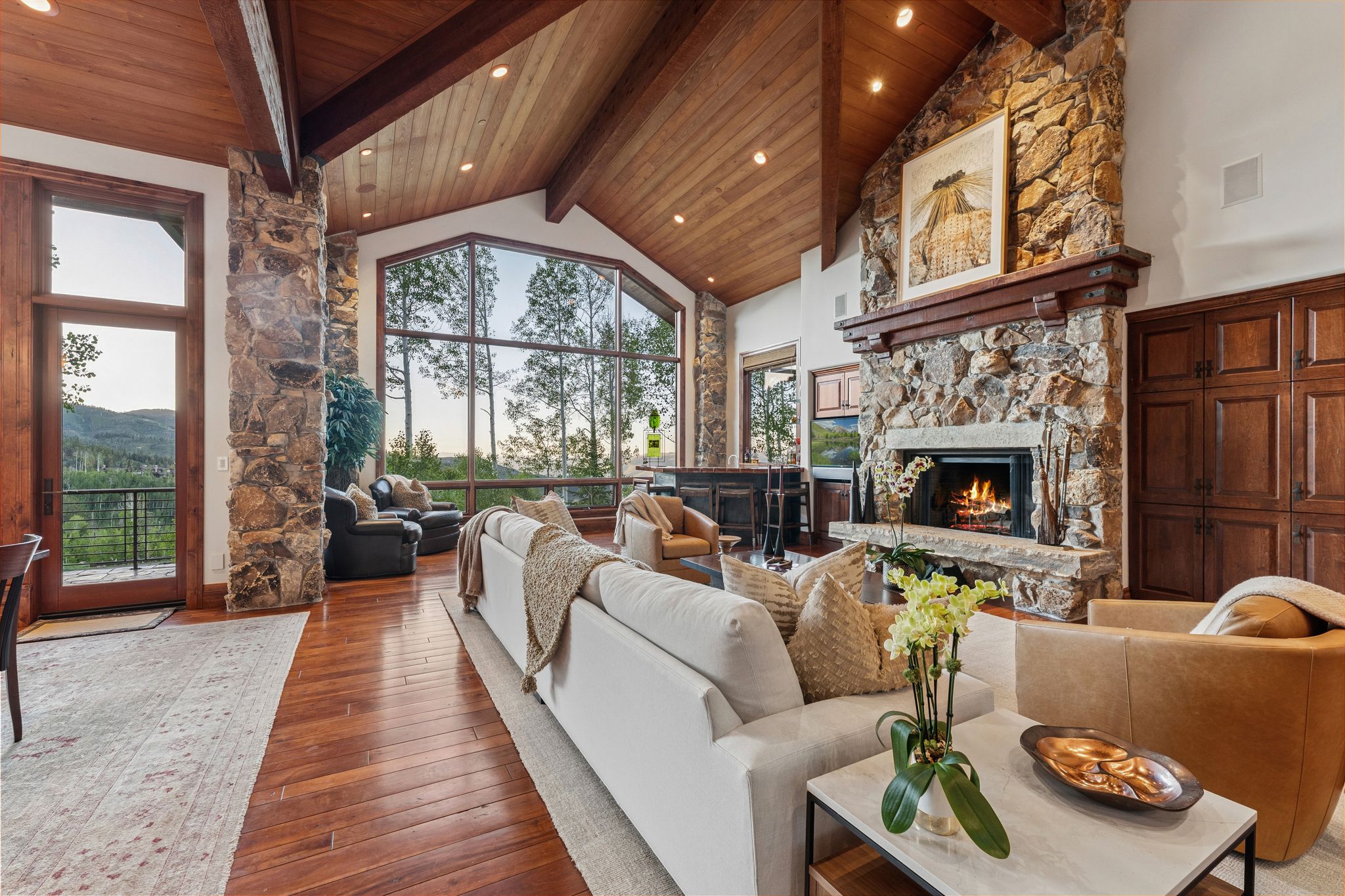 Spacious living area featuring a vaulted wood-paneled ceiling with exposed beams, a large stone fireplace with a substantial wood mantel, a wall of windows offering expansive views, rich wood-finish flooring, and built-in wood cabinetry
