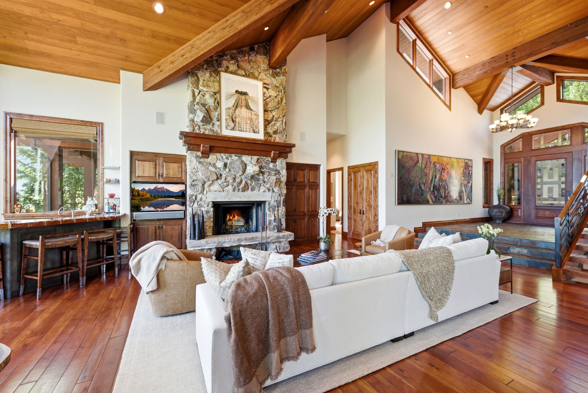 Spacious living area featuring a stone fireplace with a substantial wood mantel, vaulted wood-paneled ceilings with exposed wood beams, and rich wood-finish flooring