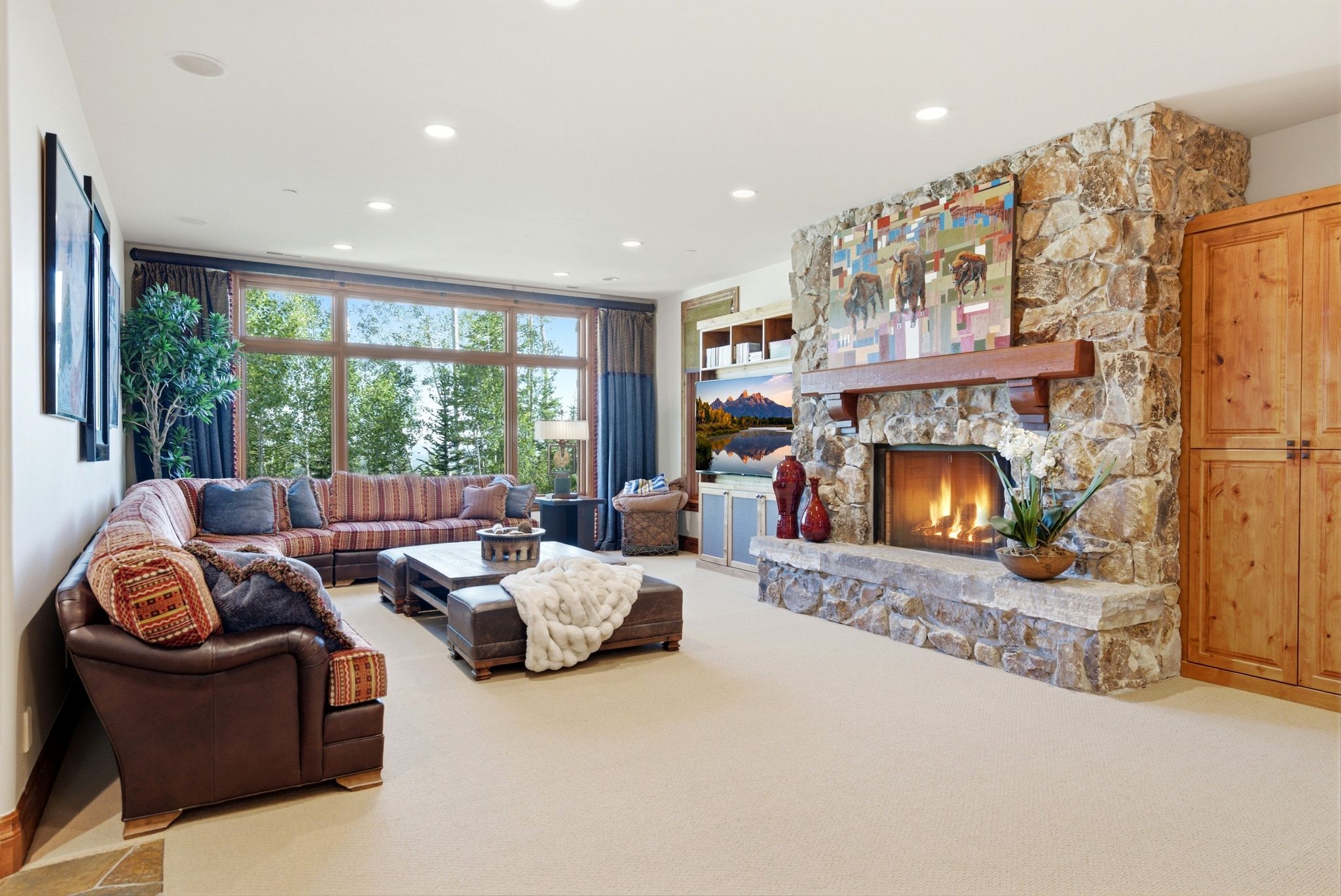 Spacious living area featuring a stone-clad fireplace with a substantial timber mantel, large picture windows, recessed ceiling lighting, and built-in wood cabinetry
