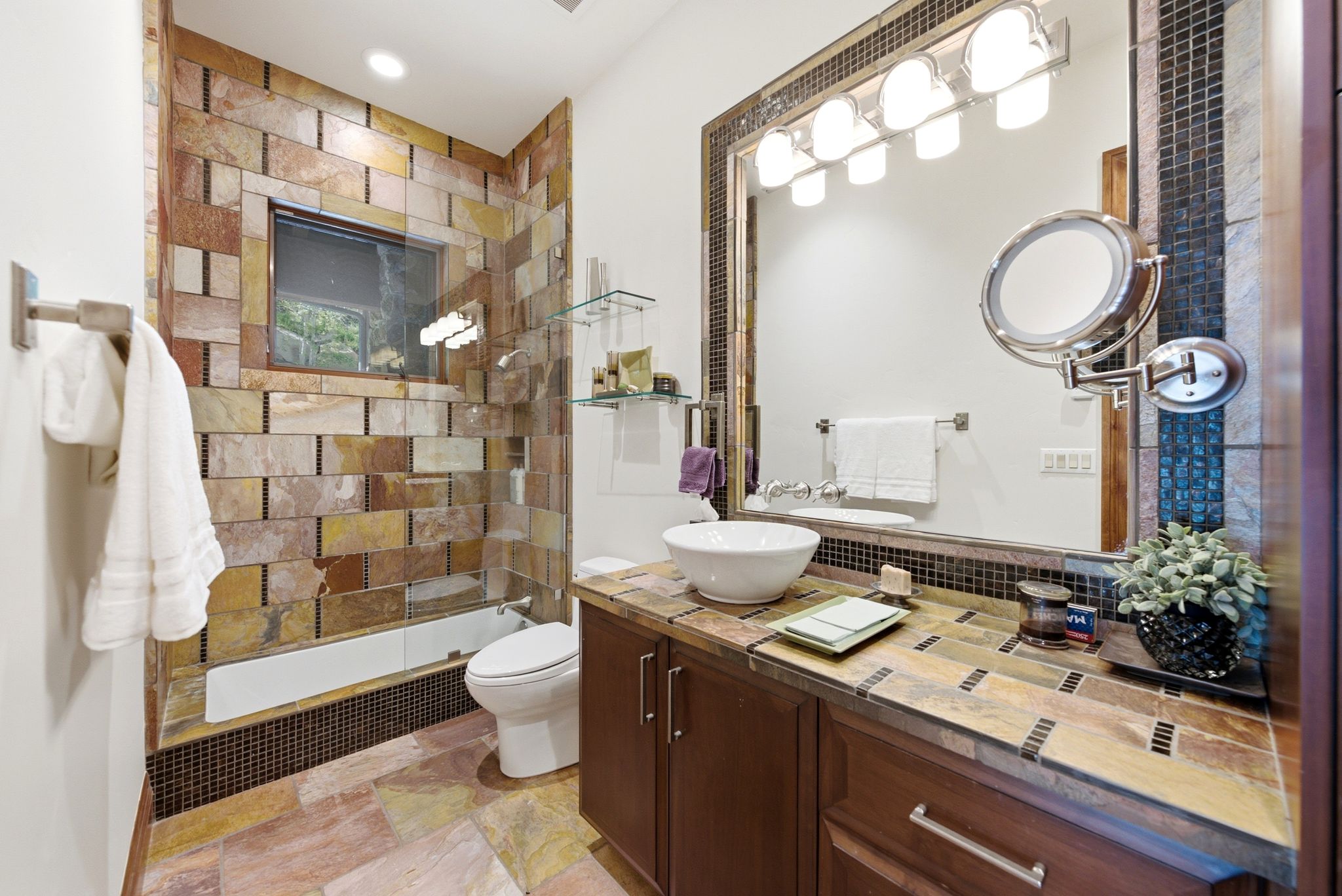 Bathroom featuring extensive natural stone tilework, a vessel sink, dark wood-finish cabinetry, and a full-width vanity mirror with integrated lighting