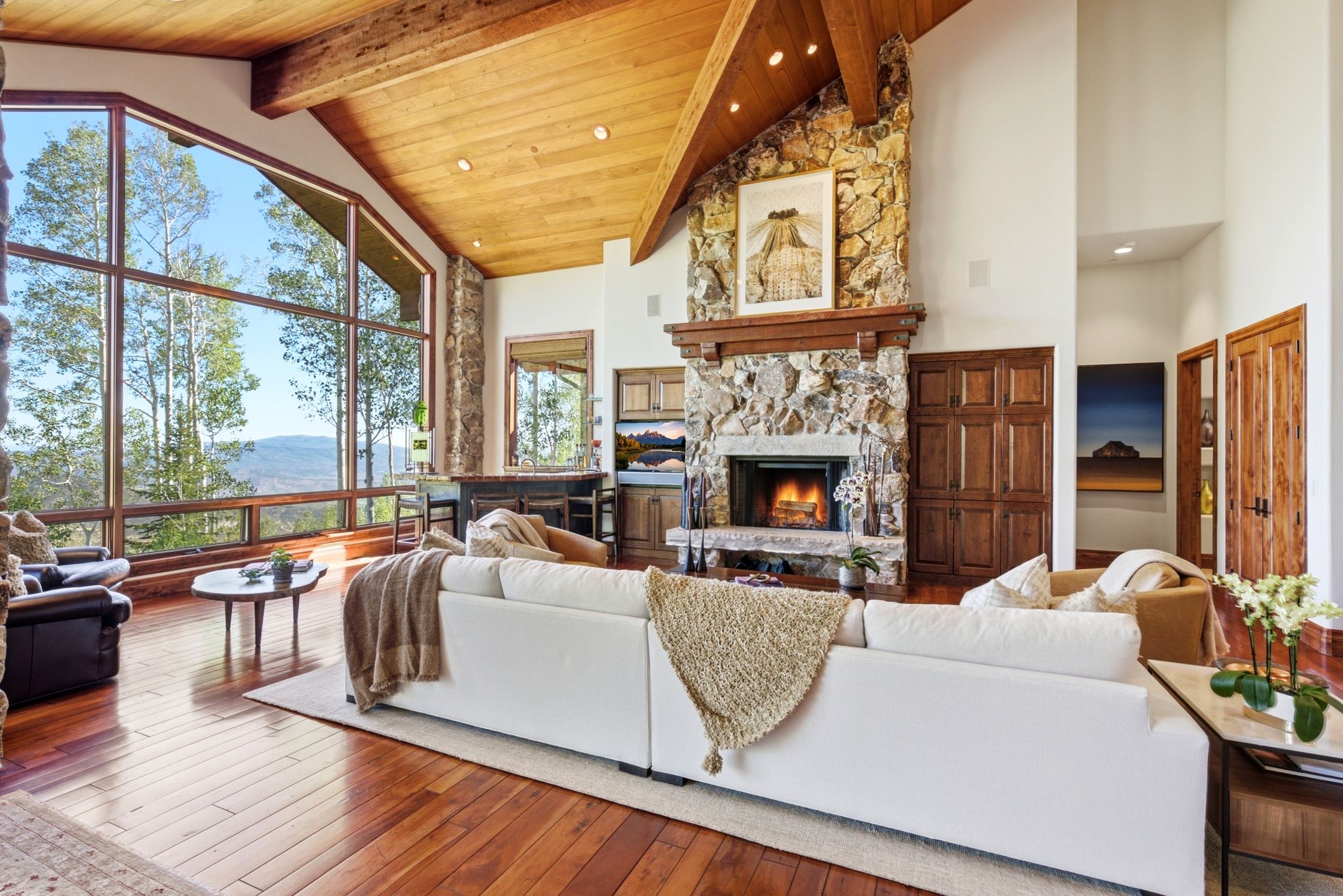 Great room featuring a vaulted wood-beam ceiling, floor-to-ceiling windows, a stone-clad fireplace with a wood mantel, wood-finish flooring, and built-in bar cabinetry