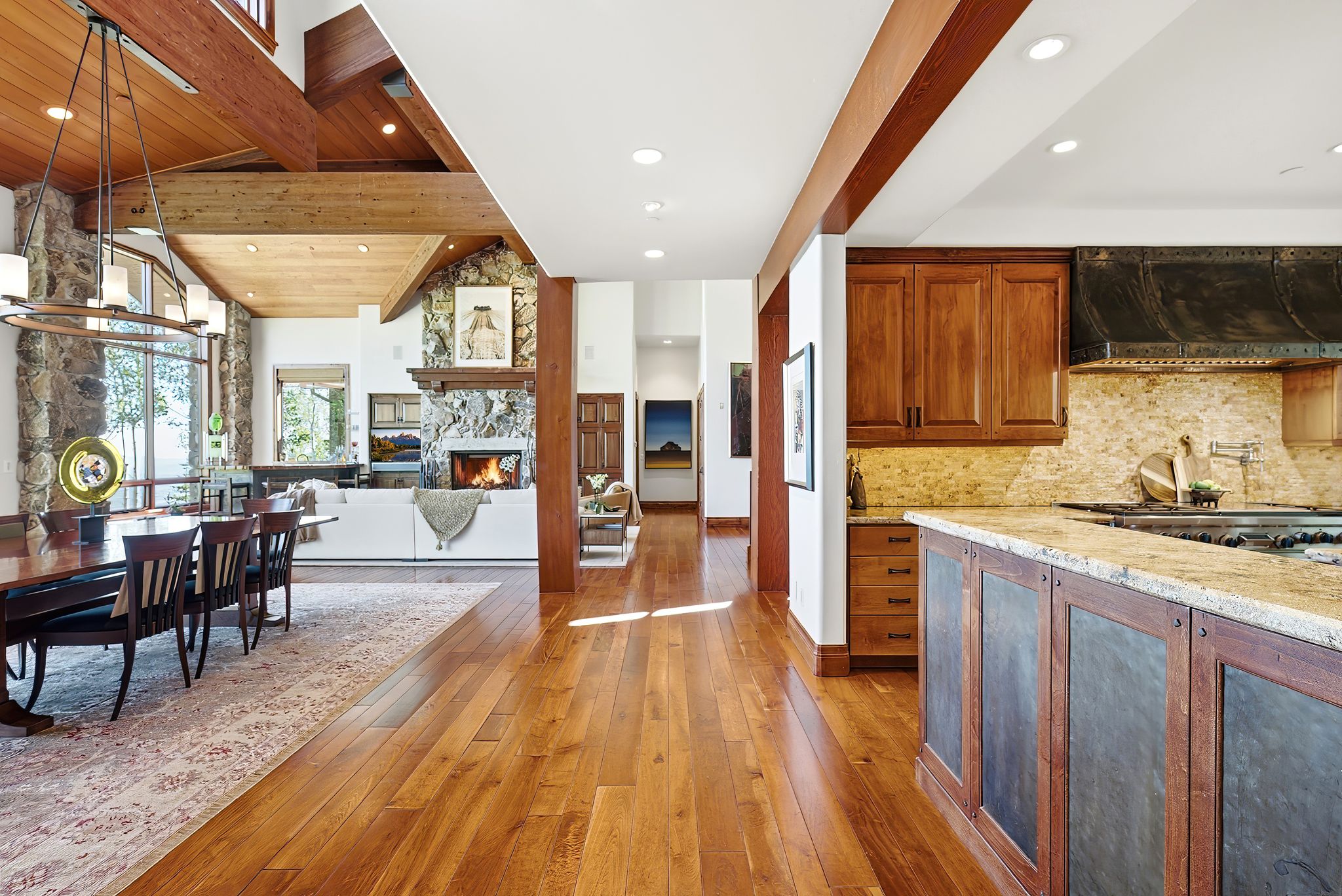 Open-concept living space featuring wood-finish flooring, exposed wood beams, a stone-clad fireplace with a wood mantle, and large windows