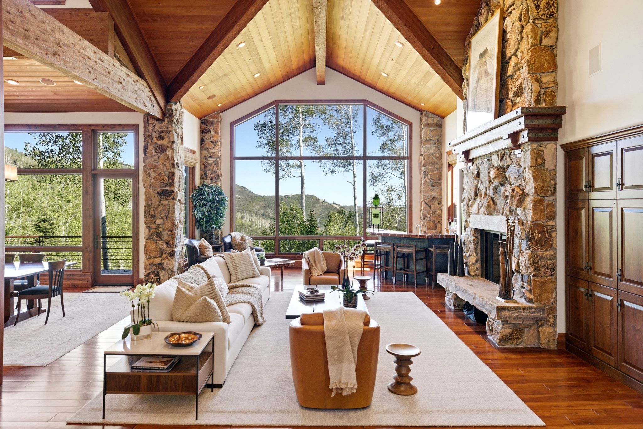 Vaulted great room with exposed wood beams, expansive floor-to-ceiling windows, natural stone fireplace, wood-finish flooring, and built-in cabinetry