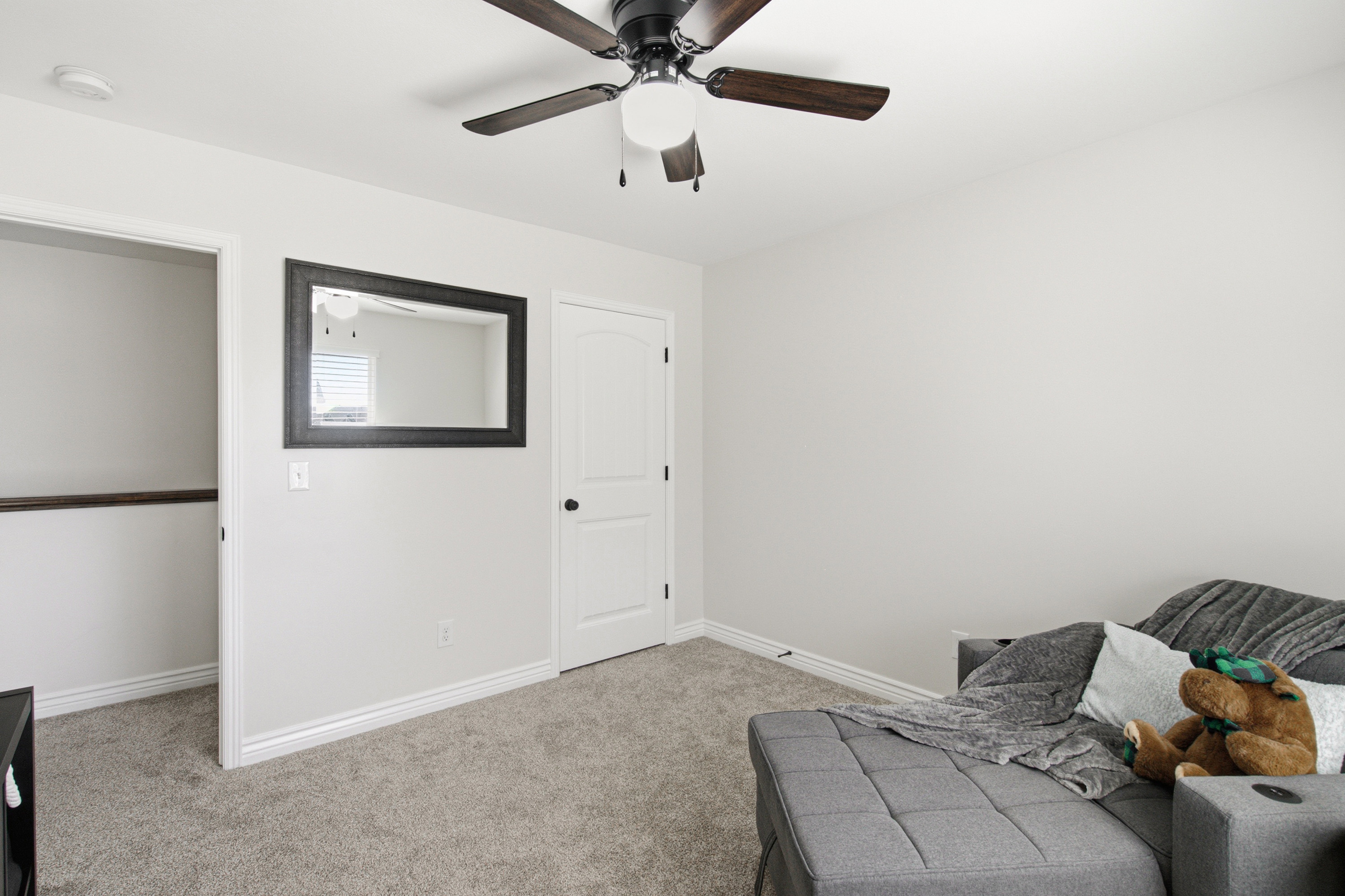 Carpeted room with a ceiling fan featuring wood-finish blades, a white paneled door with black hardware, and crisp white baseboards