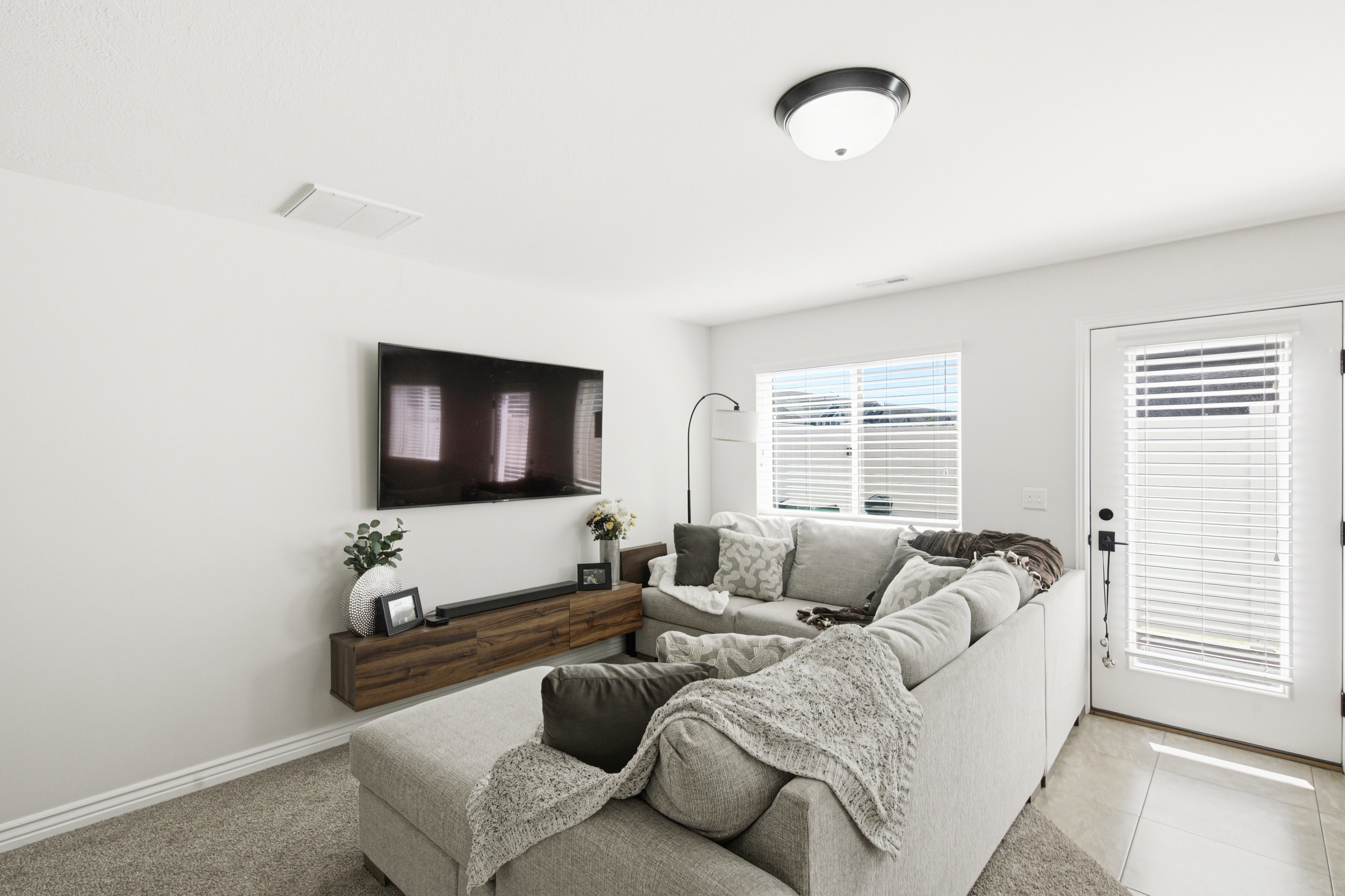 Living area featuring a wall-mounted television, a floating wood-finish media console, a window with blinds, and a door with an integrated blind, all under a flush mount ceiling light