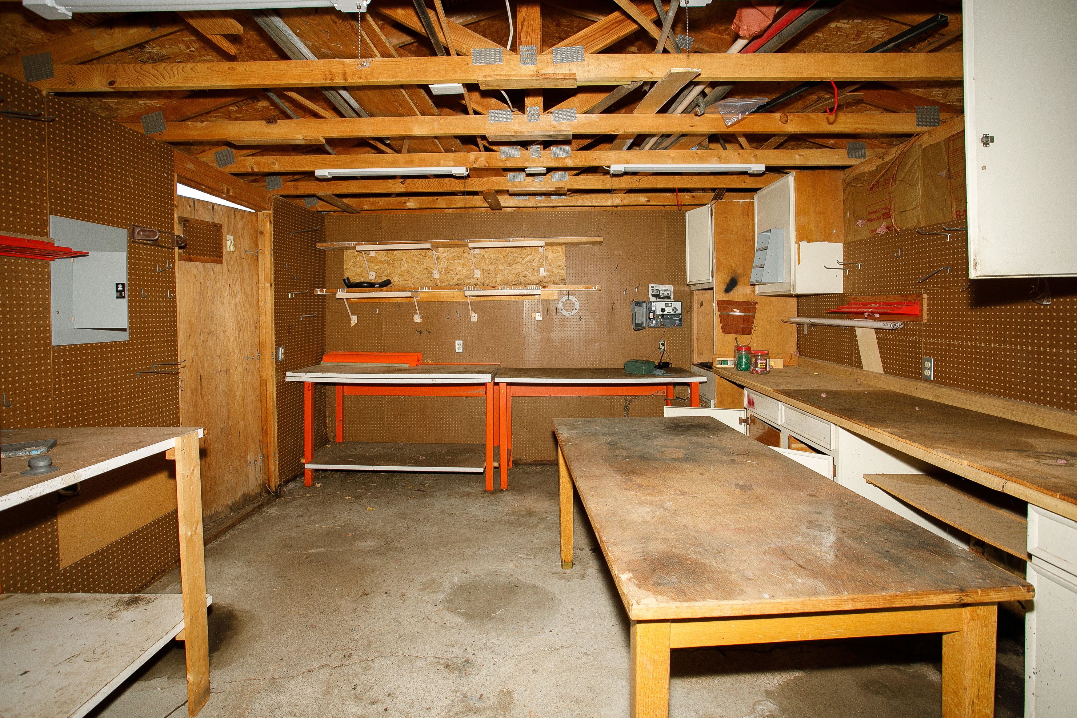 Utility space featuring exposed wood rafter ceiling, multiple fluorescent light fixtures, extensive pegboard wall storage, integrated shelving, and built-in cabinetry