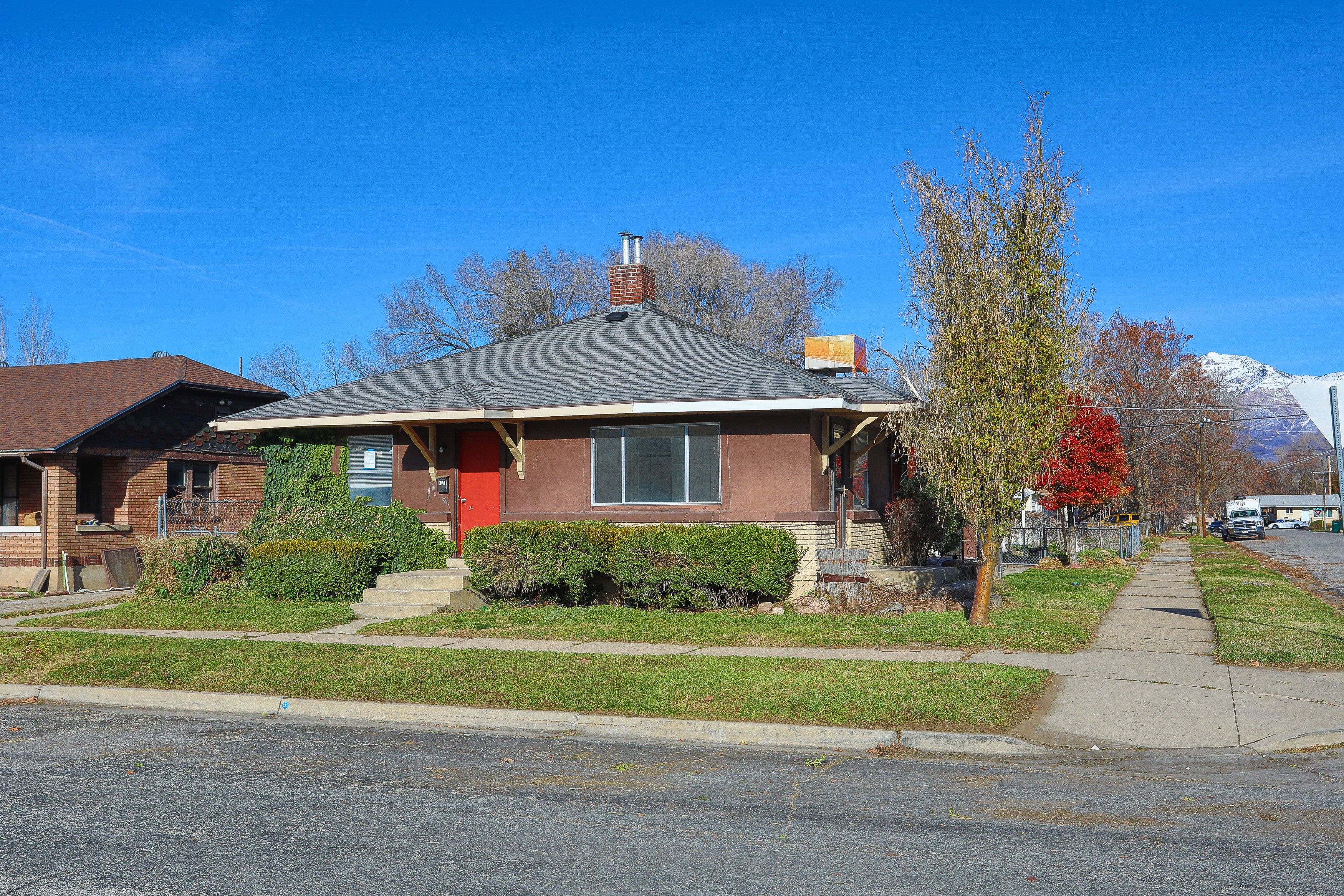 Ranch-style residence featuring a brown facade, a red entry door, a gray shingle roof, and a visible chimney