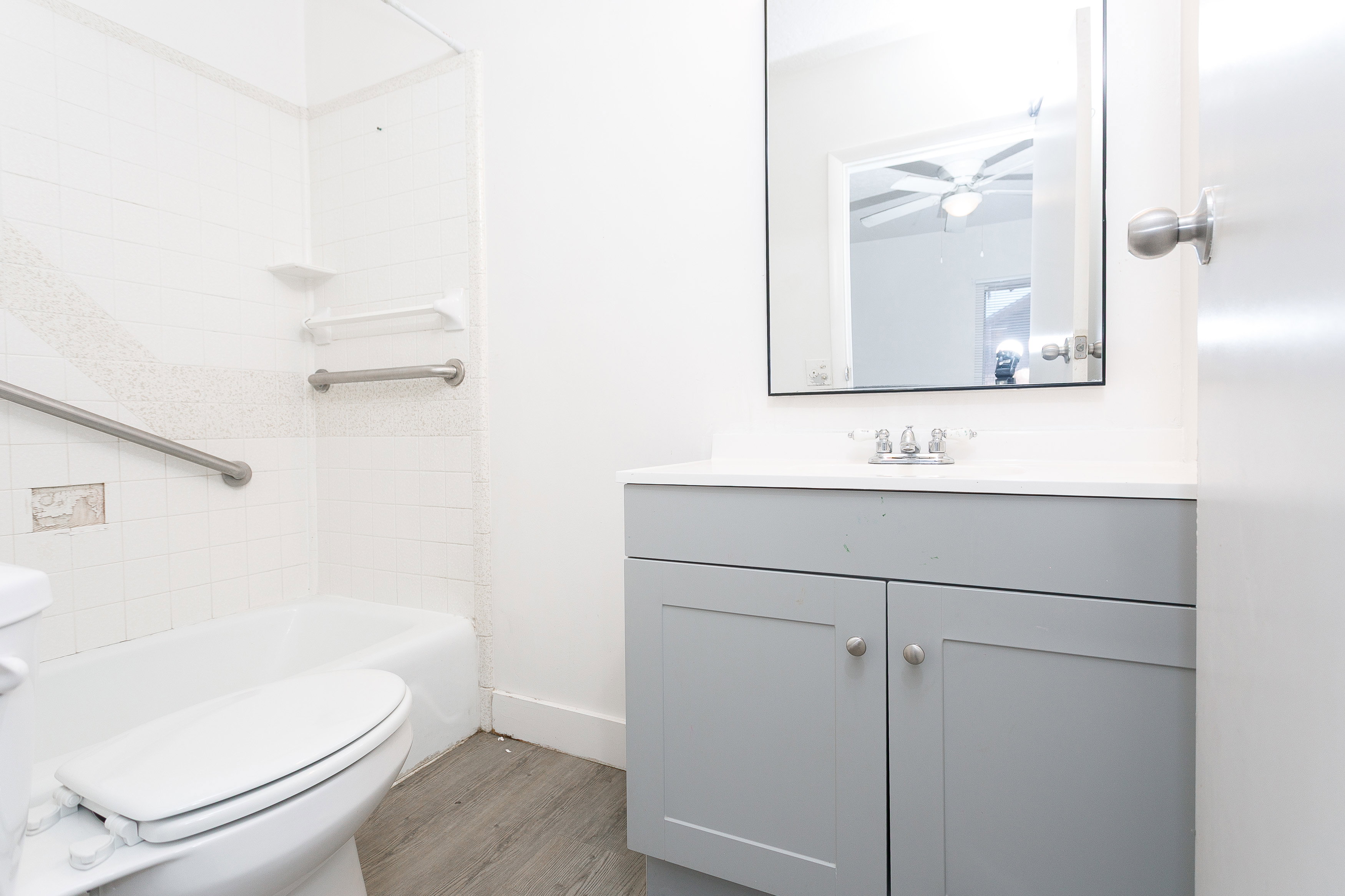Bathroom featuring a white bathtub, tiled shower surround with grab bars, gray wood-finish vanity with a white countertop, and a wall-mounted mirror