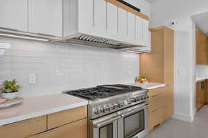 Gourmet kitchen featuring a professional-grade stainless steel range with griddle, white subway tile backsplash, white countertops, natural wood-finish lower cabinetry, and white upper cabinetry with integrated lighting