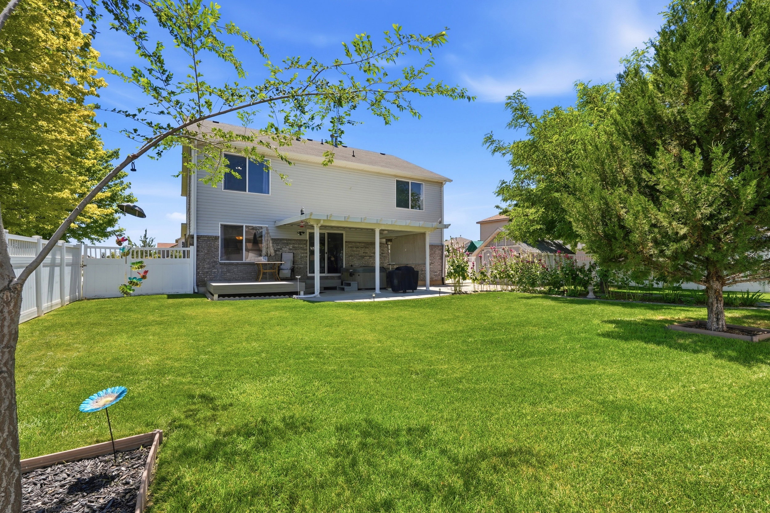 Spacious backyard featuring a large lawn, mature trees, white picket fencing, and a covered patio area