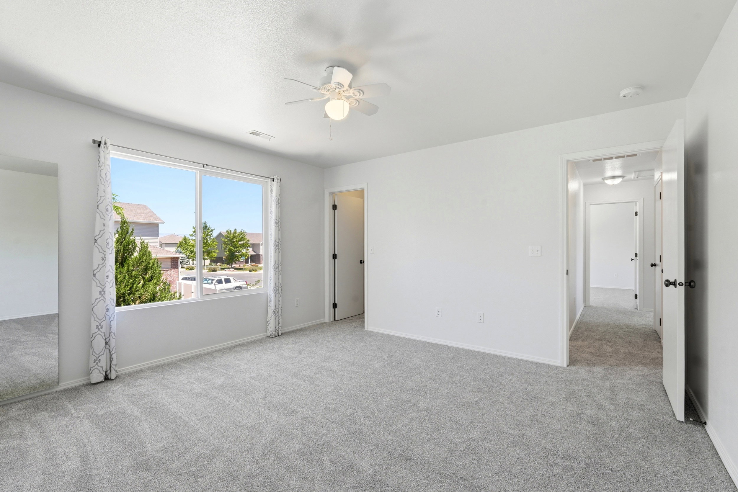 Spacious room featuring light gray carpet, white walls, a large window with a neighborhood perspective, and an overhead ceiling fan