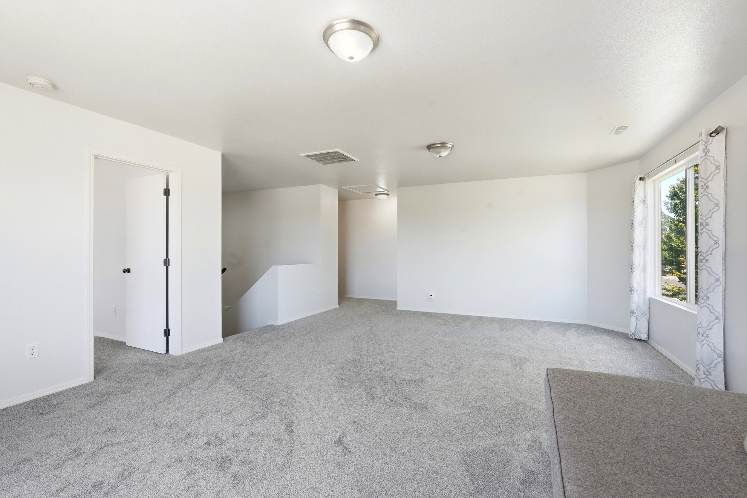 Spacious room featuring light gray carpet flooring, white walls, multiple flush-mount ceiling lights, a large window, and an open doorway