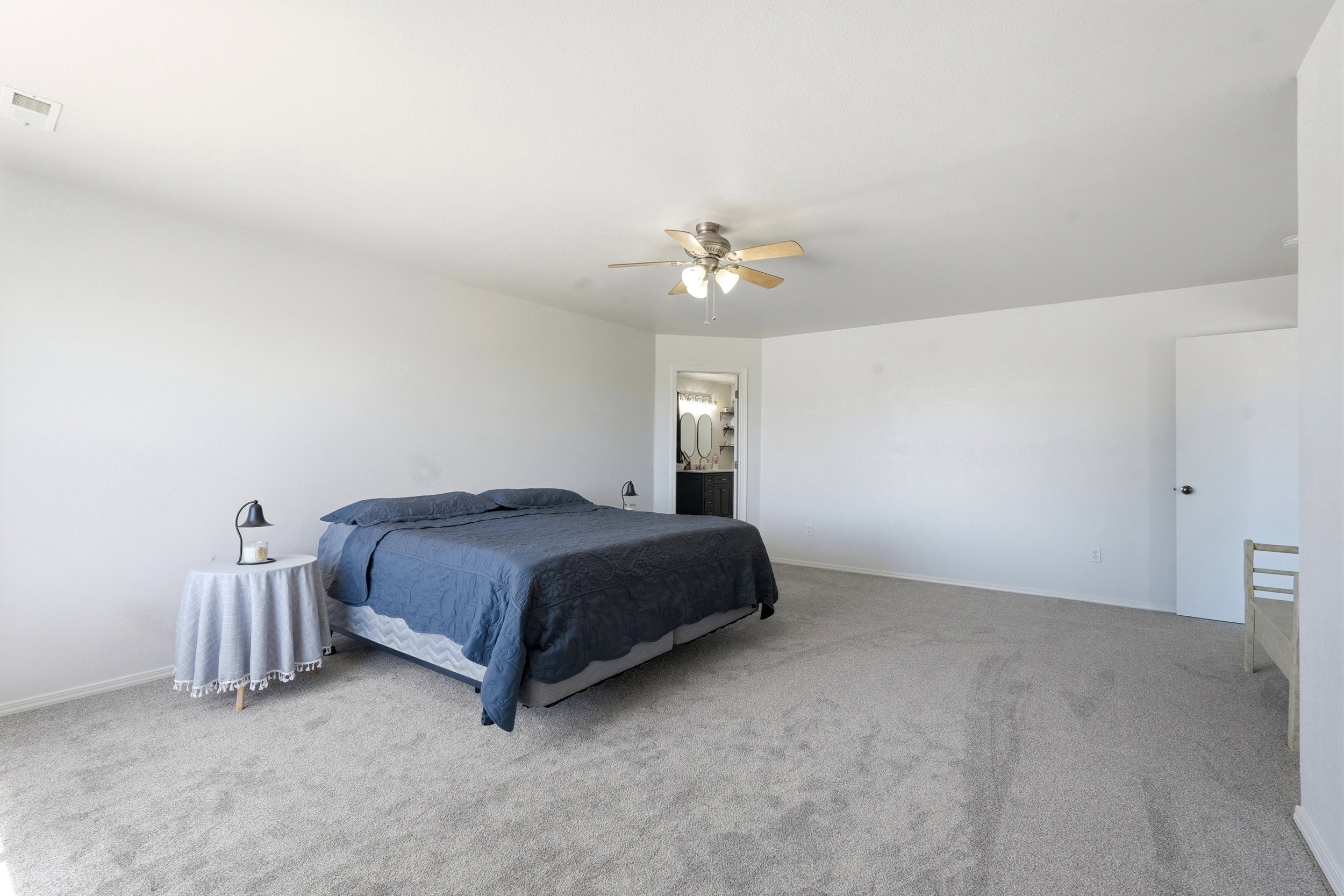 Spacious bedroom featuring light neutral carpeting, a ceiling fan with light kit, and a doorway opening to an en-suite bathroom with a double vanity