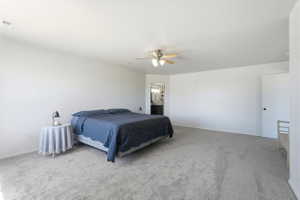 Spacious bedroom featuring light neutral carpeting, a ceiling fan with light kit, and a doorway opening to an en-suite bathroom with a double vanity