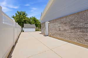 Paved side yard featuring a white vinyl privacy fence, a brick exterior wall with a service door, and a detached storage shed with a shingle roof