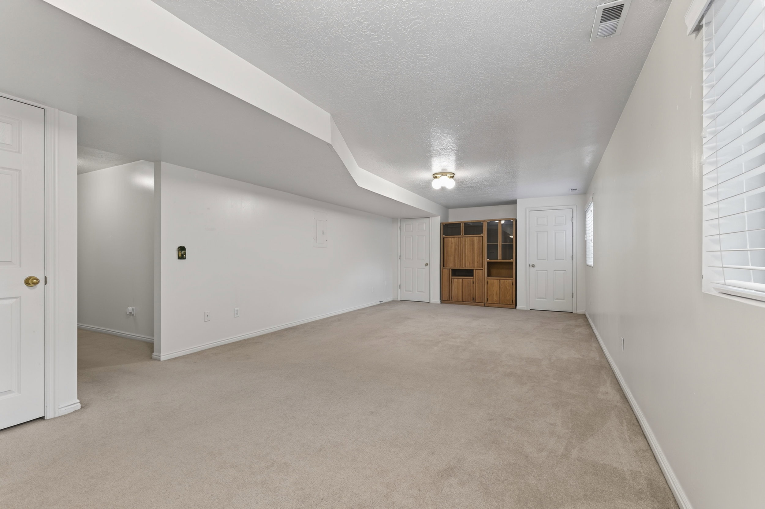 Spacious lower-level room featuring light-toned carpeting, white painted walls, and a built-in wood-finish cabinet with glass doors