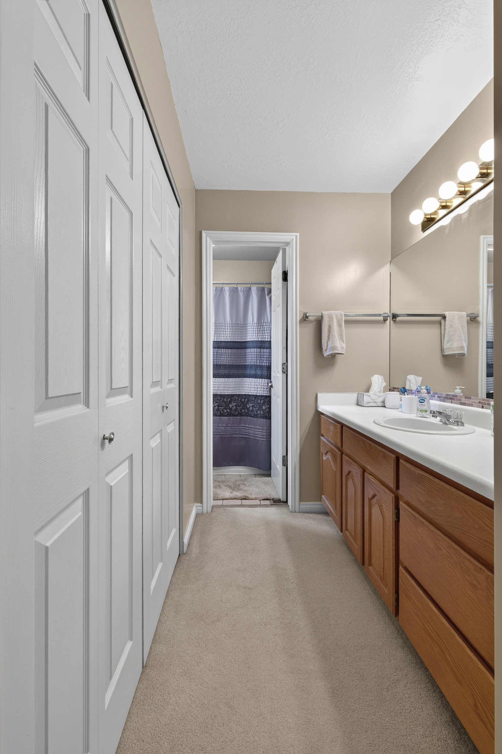 Bathroom featuring a light beige carpet, a long wood-finish vanity with a white countertop and integrated sink, a wall-mounted mirror, and a multi-bulb light fixture