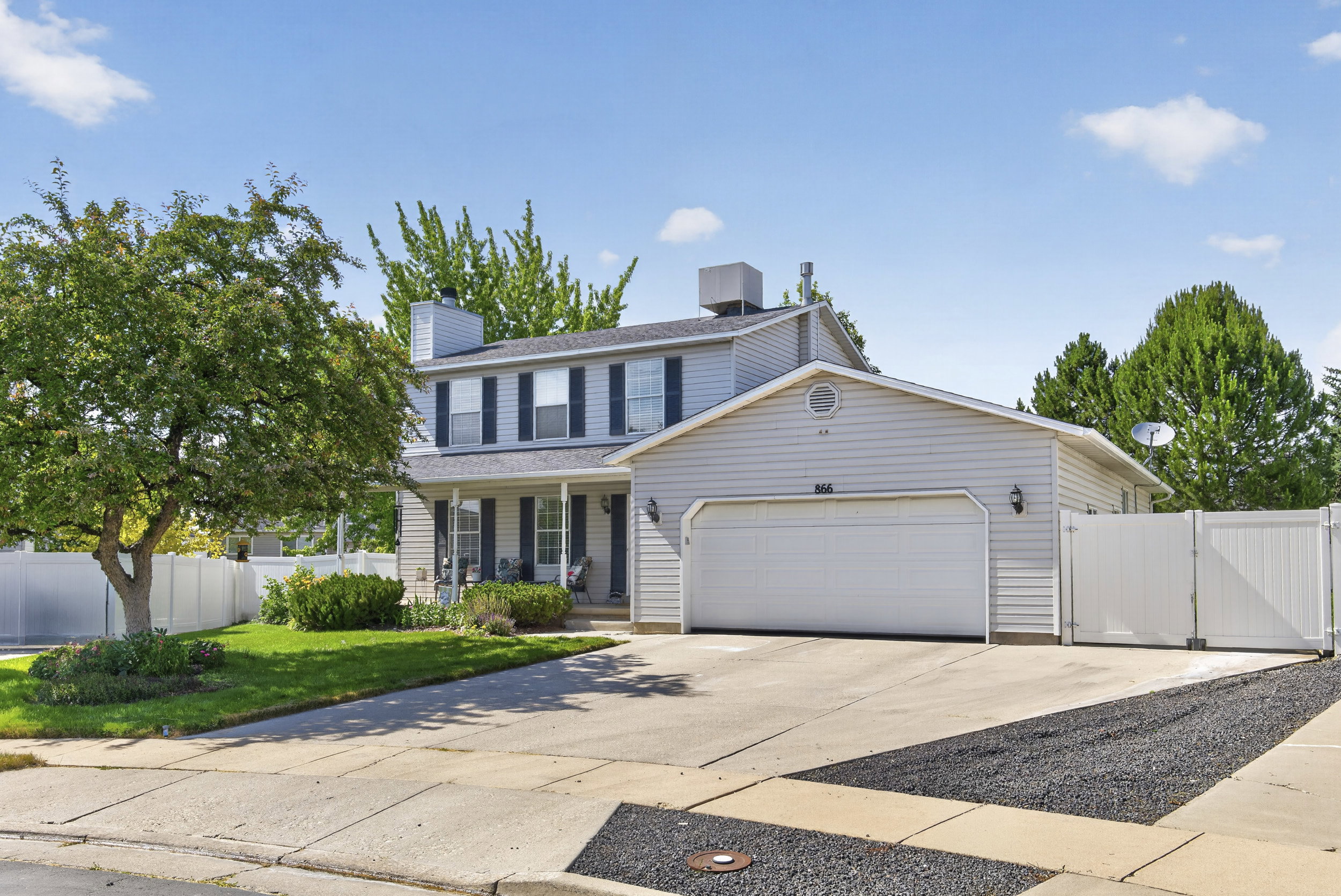 Light-colored siding exterior with dark shutters, covered front porch, attached two-car garage, concrete driveway, and white privacy fencing