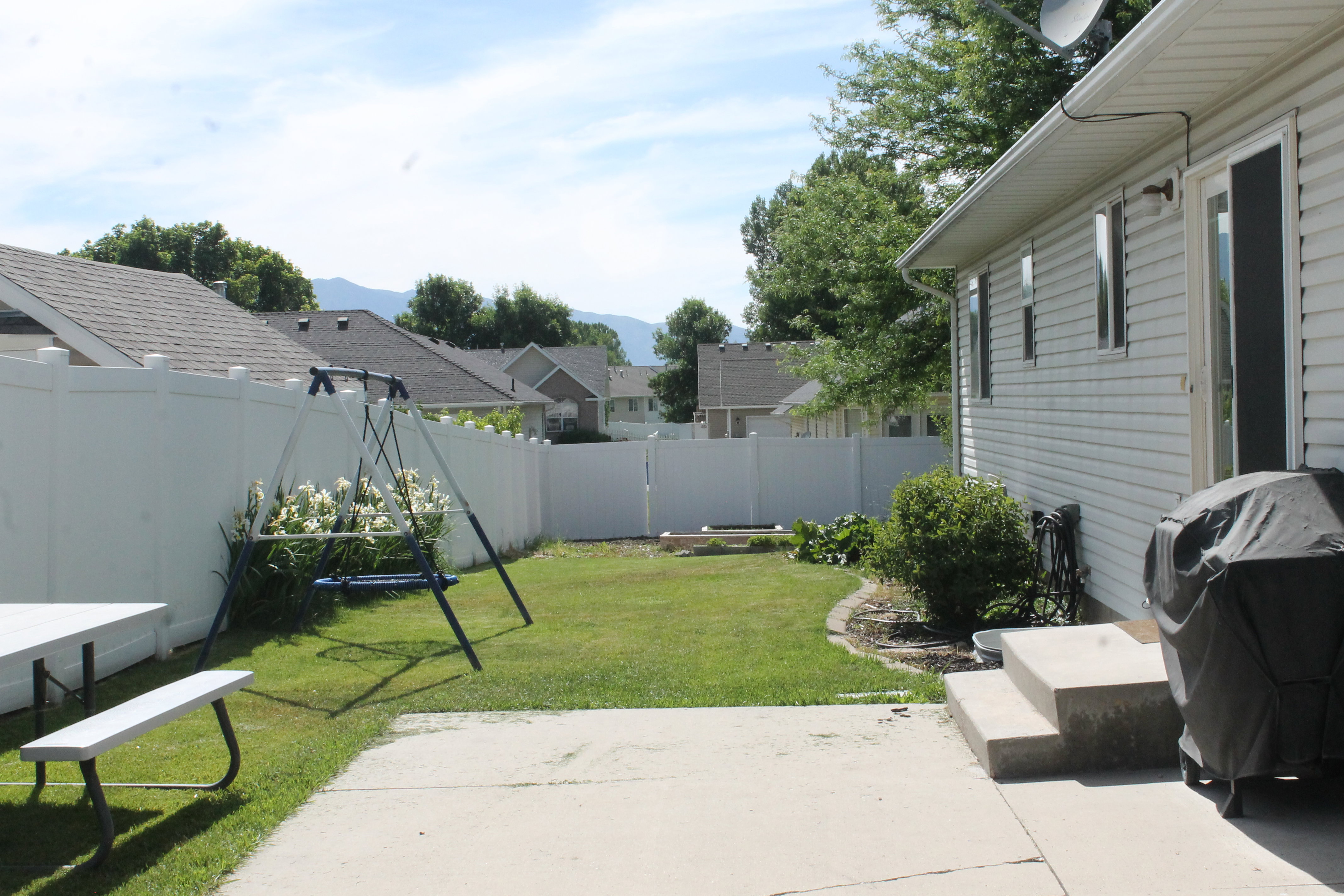 Expansive backyard featuring a concrete patio, mature lawn, white vinyl fencing, and flowering shrubs