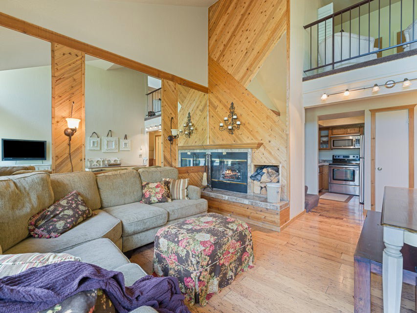 Living area featuring a stone fireplace with wood-finish paneling, wood-finish flooring, a kitchen with stainless steel appliances, and an upper-level balcony with metal railing