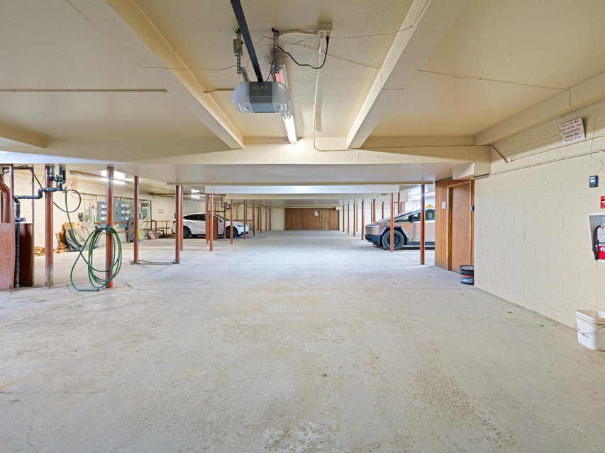 Spacious concrete-floored garage featuring exposed ceiling beams, fluorescent lighting, and a garage door opener system