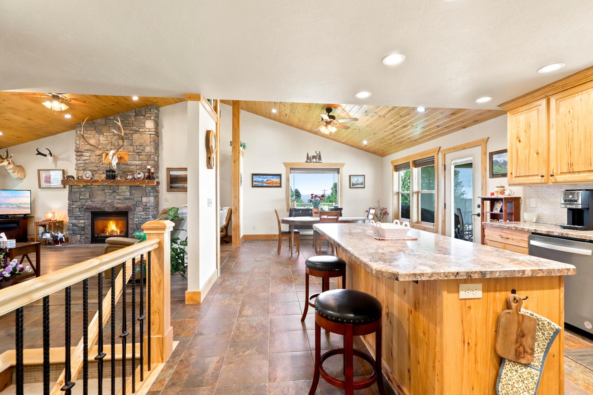 Open-concept kitchen and living area featuring a stone fireplace, wood-finish vaulted ceilings, a large kitchen island with counter seating, and light wood cabinetry with granite-style countertops