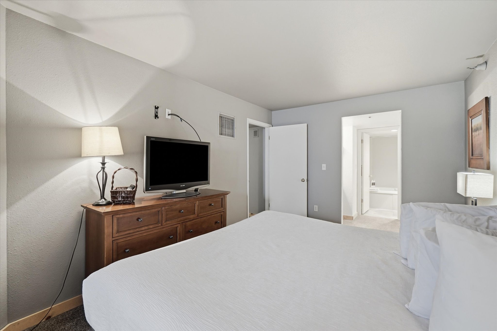 Spacious bedroom featuring light grey walls, carpet flooring, a built-in wall vent, and a hallway leading to an additional room with a built-in bathtub