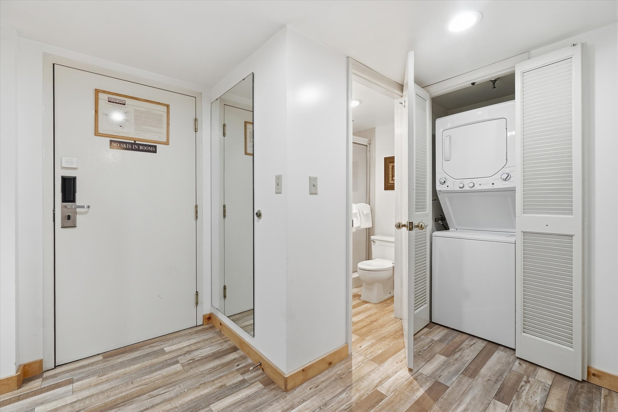 Wood-finish flooring throughout, featuring a stacked washer and dryer in a louvered bi-fold door closet, and a partial view of a bathroom with a white toilet