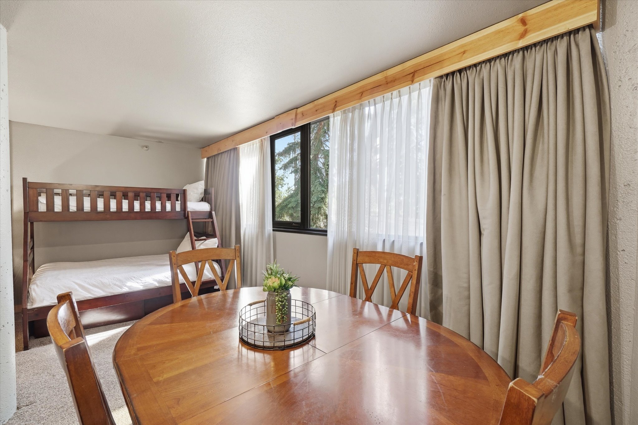 Open concept room featuring a wood-finish dining table and chairs, bunk beds, and large windows with privacy drapes