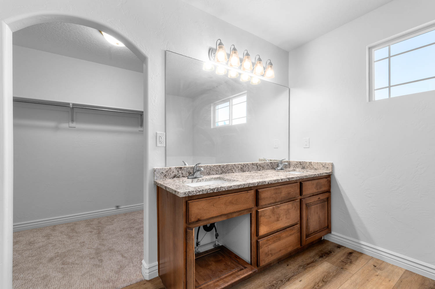 Bathroom featuring a double vanity with granite countertop and dual sinks, wood-finish flooring, a large mirror, and an arched doorway leading to a carpeted closet with shelving