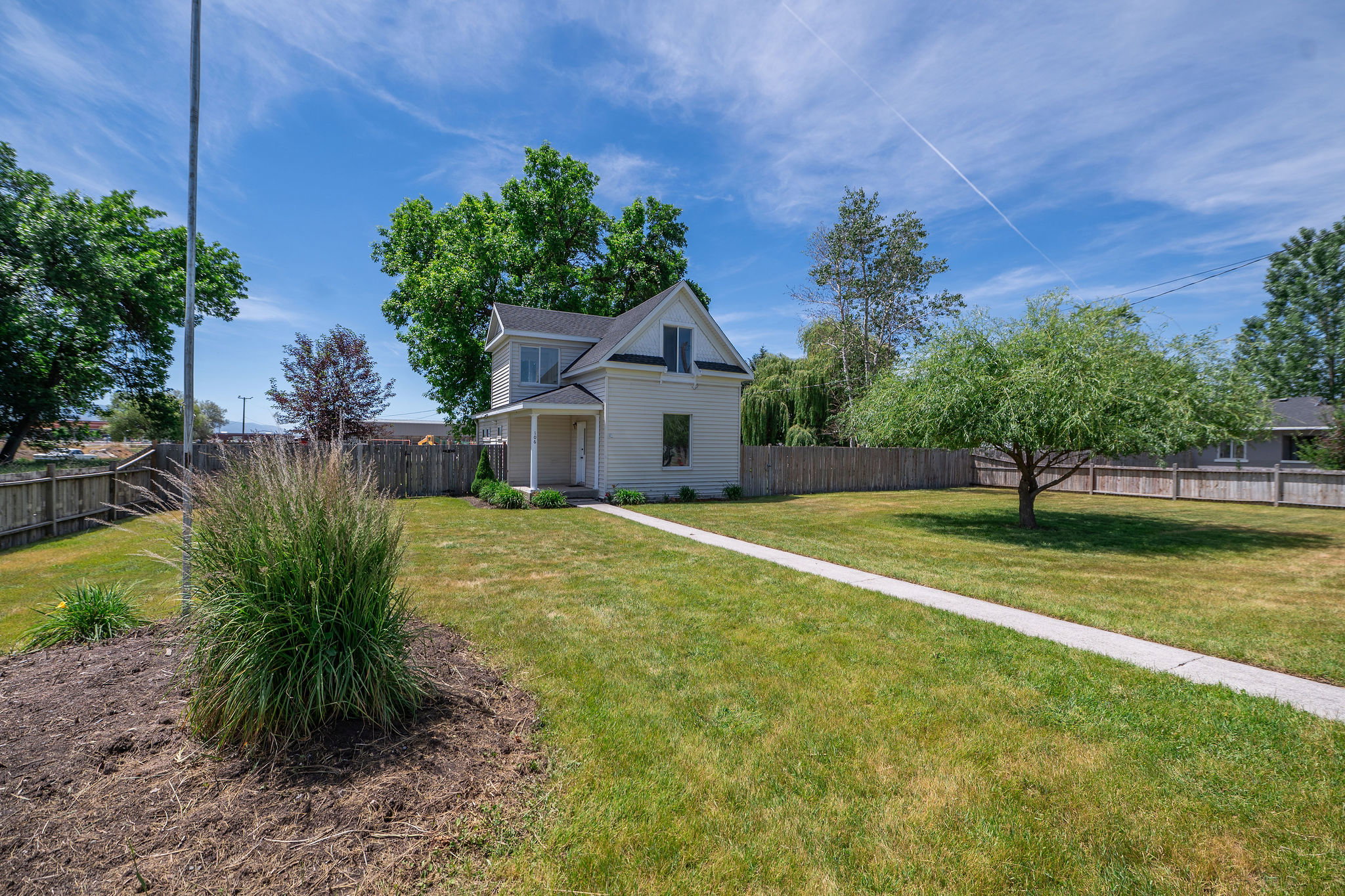 Cream-colored two-story residence with a covered porch and dormer window, situated on a grassy lot with mature trees and a wooden privacy fence