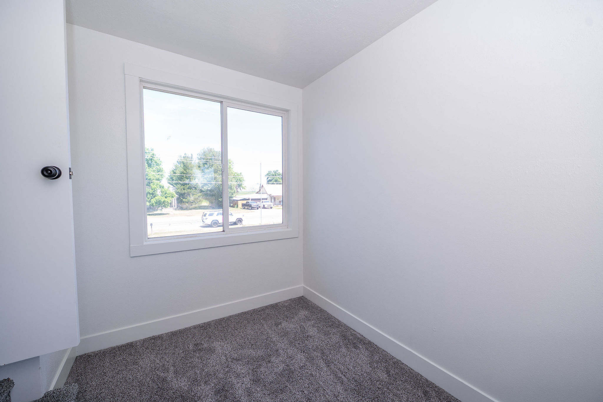 Bright room featuring a white-framed window, neutral gray carpet, white walls, and a white door with black hardware