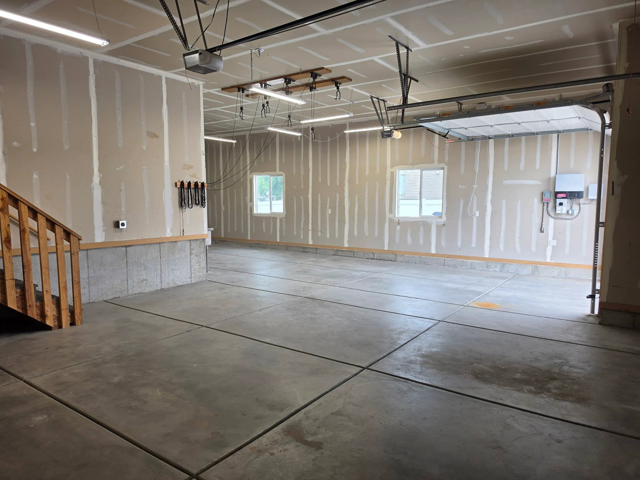 Spacious garage featuring a concrete floor, exposed drywall walls, multiple fluorescent light fixtures, two windows, and an unpainted wooden staircase