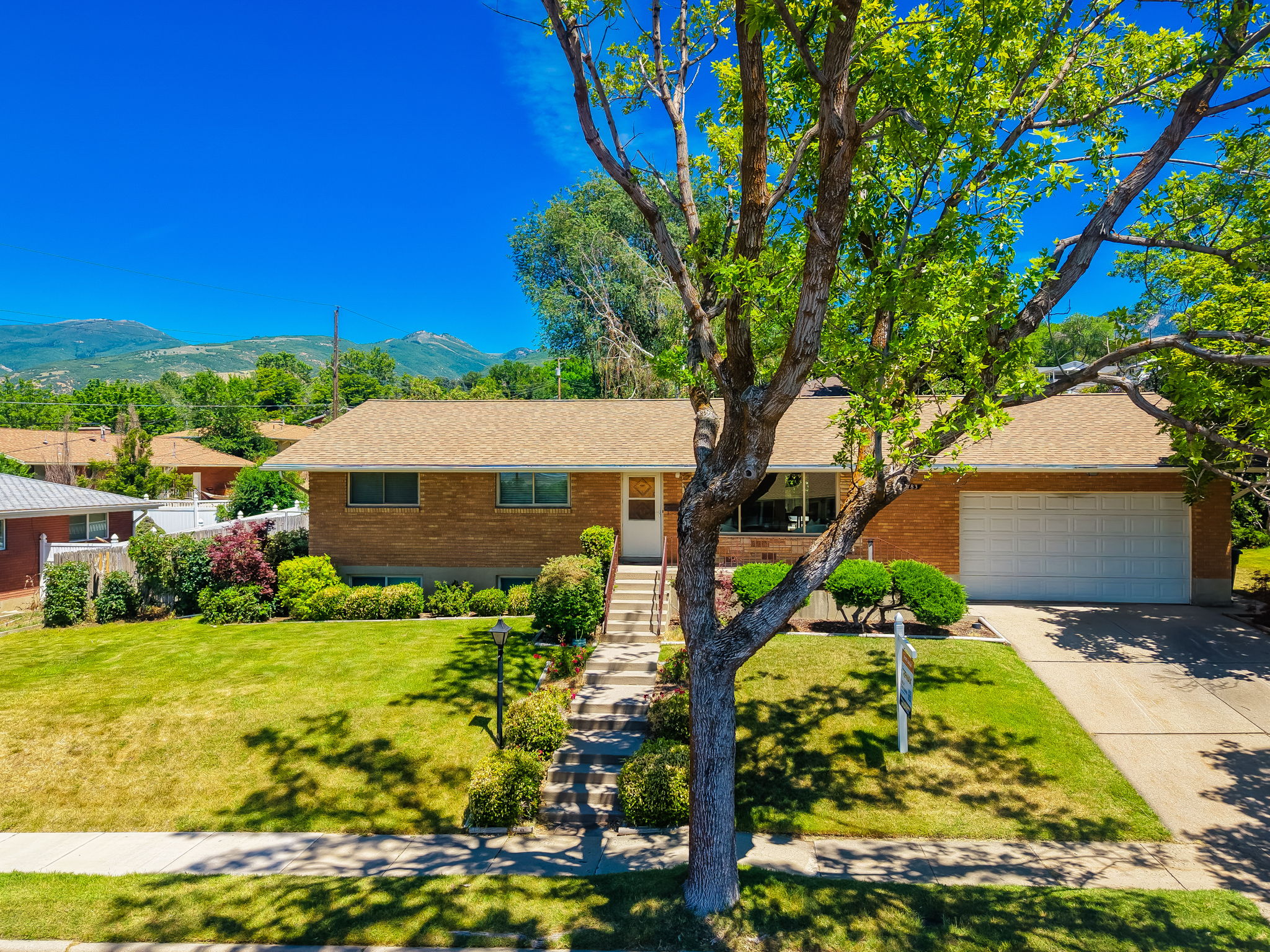Brick rambler residence featuring a front lawn, mature tree, concrete driveway, and attached garage