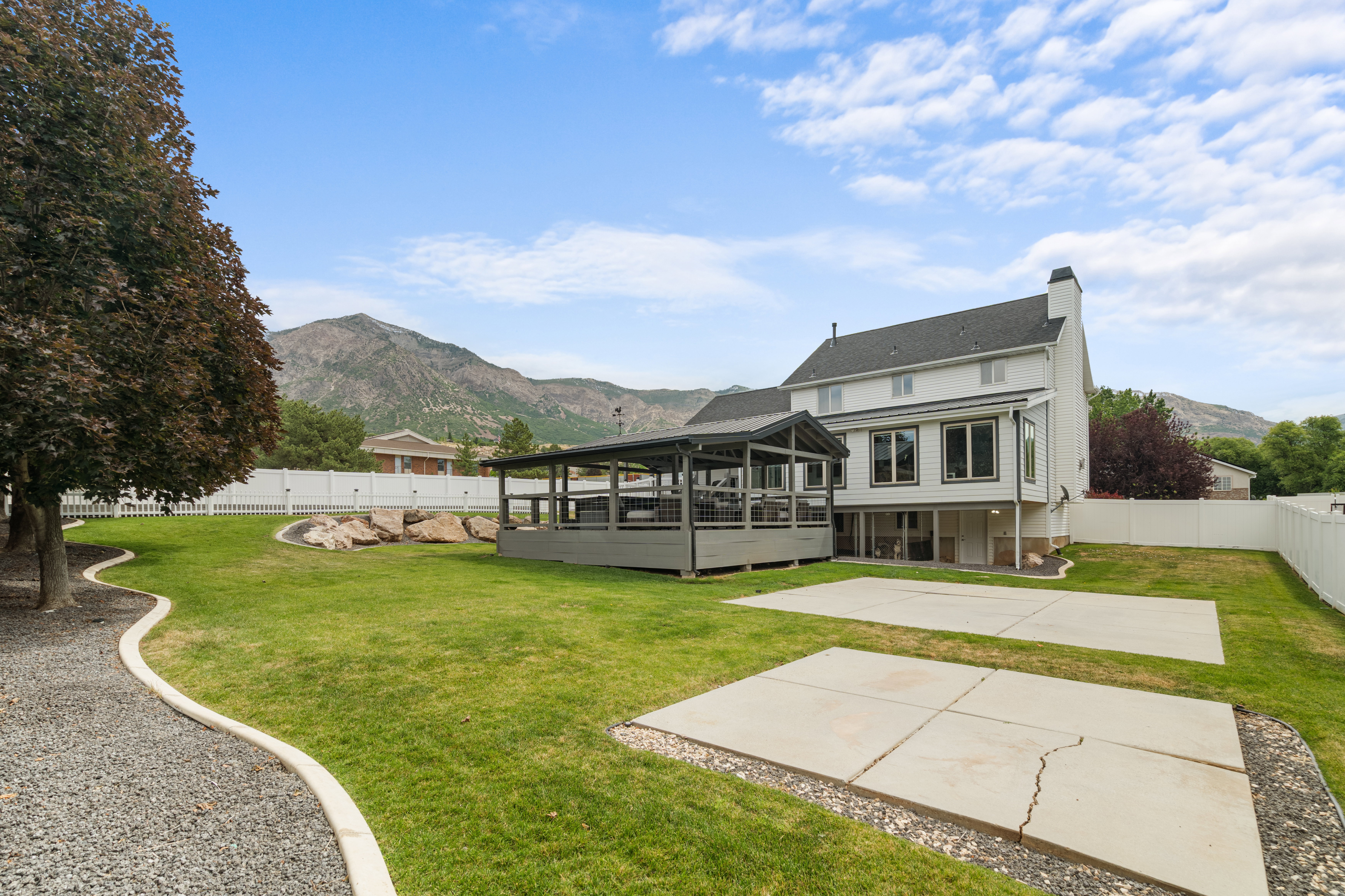 Expansive backyard featuring a covered patio deck, two concrete pad areas, mature trees, and a gravel pathway