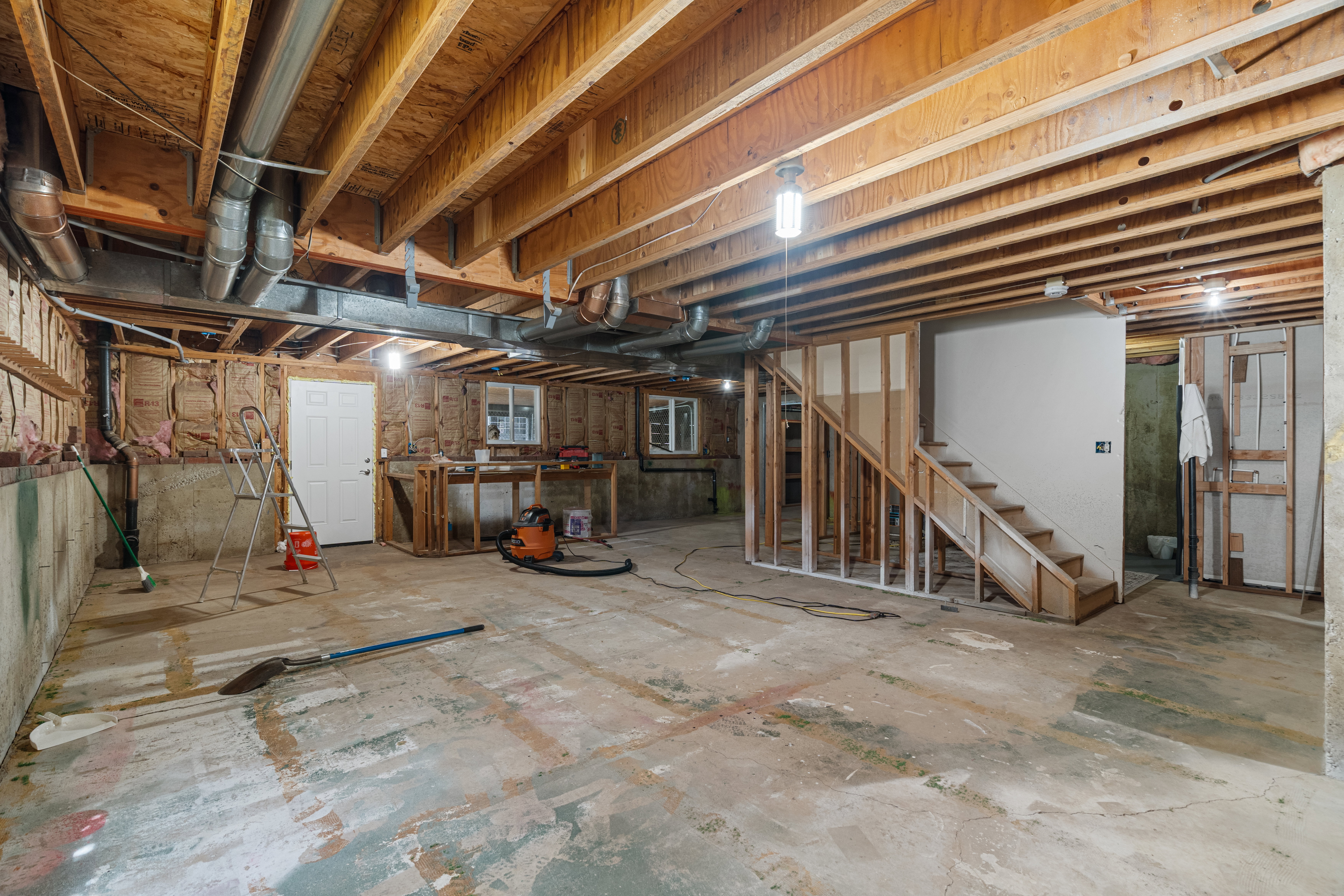 Unfinished basement space featuring exposed wooden ceiling joists, concrete flooring, partially framed walls, and an unfinished staircase